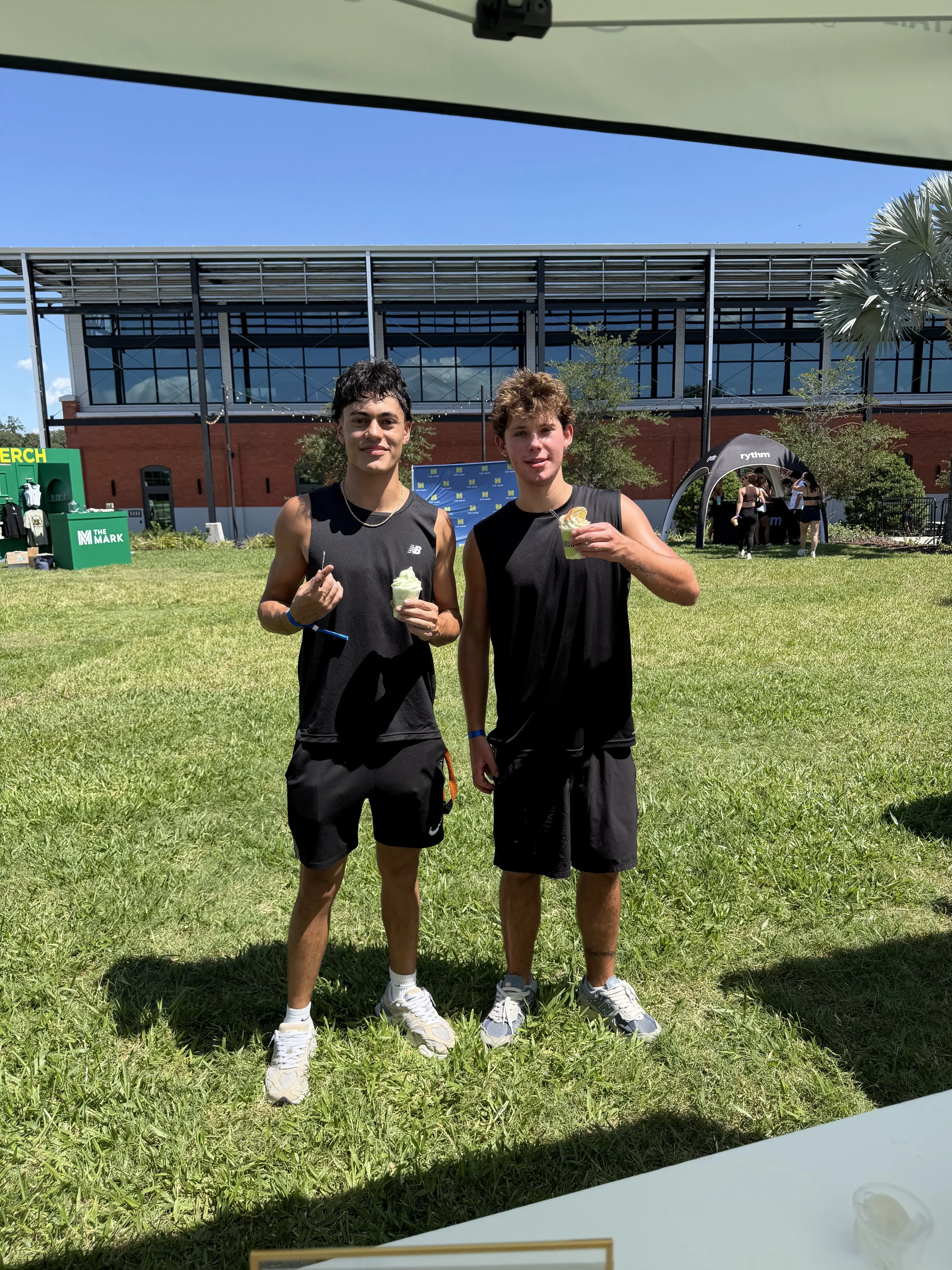 Two young men in athletic clothing standing on a grassy field, holding cups of ice cream, with a modern building, trees, and tents in the background under a clear blue sky.