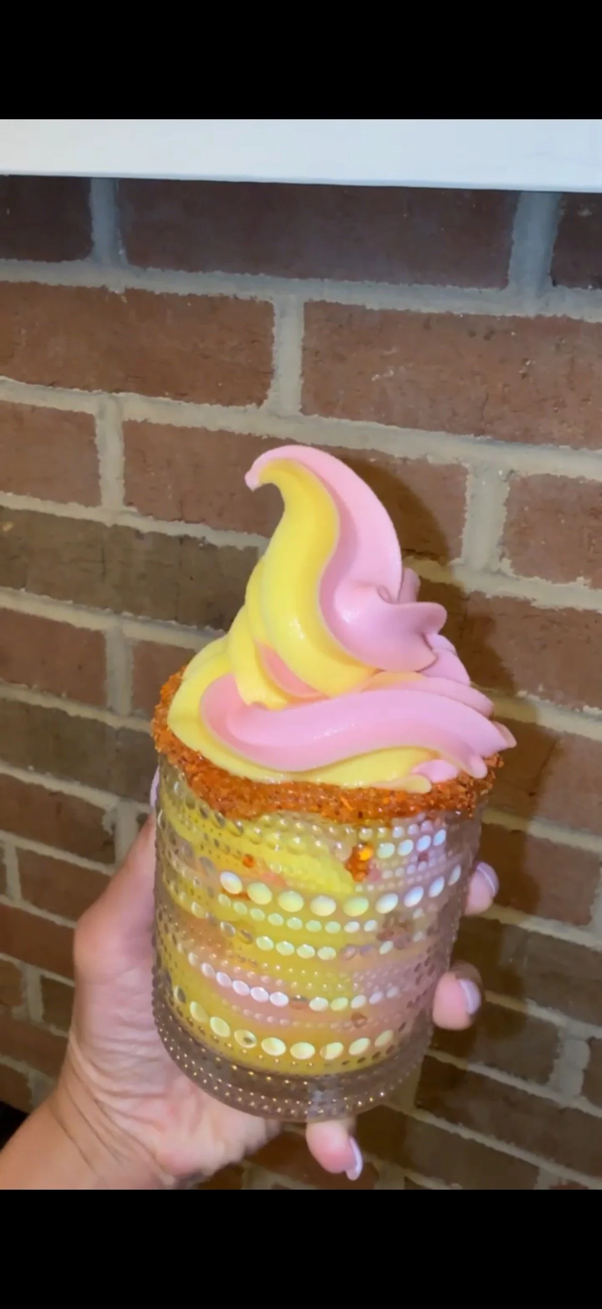 Hand holding a colorful soft-serve margarita in a decorative glass against a brick wall.