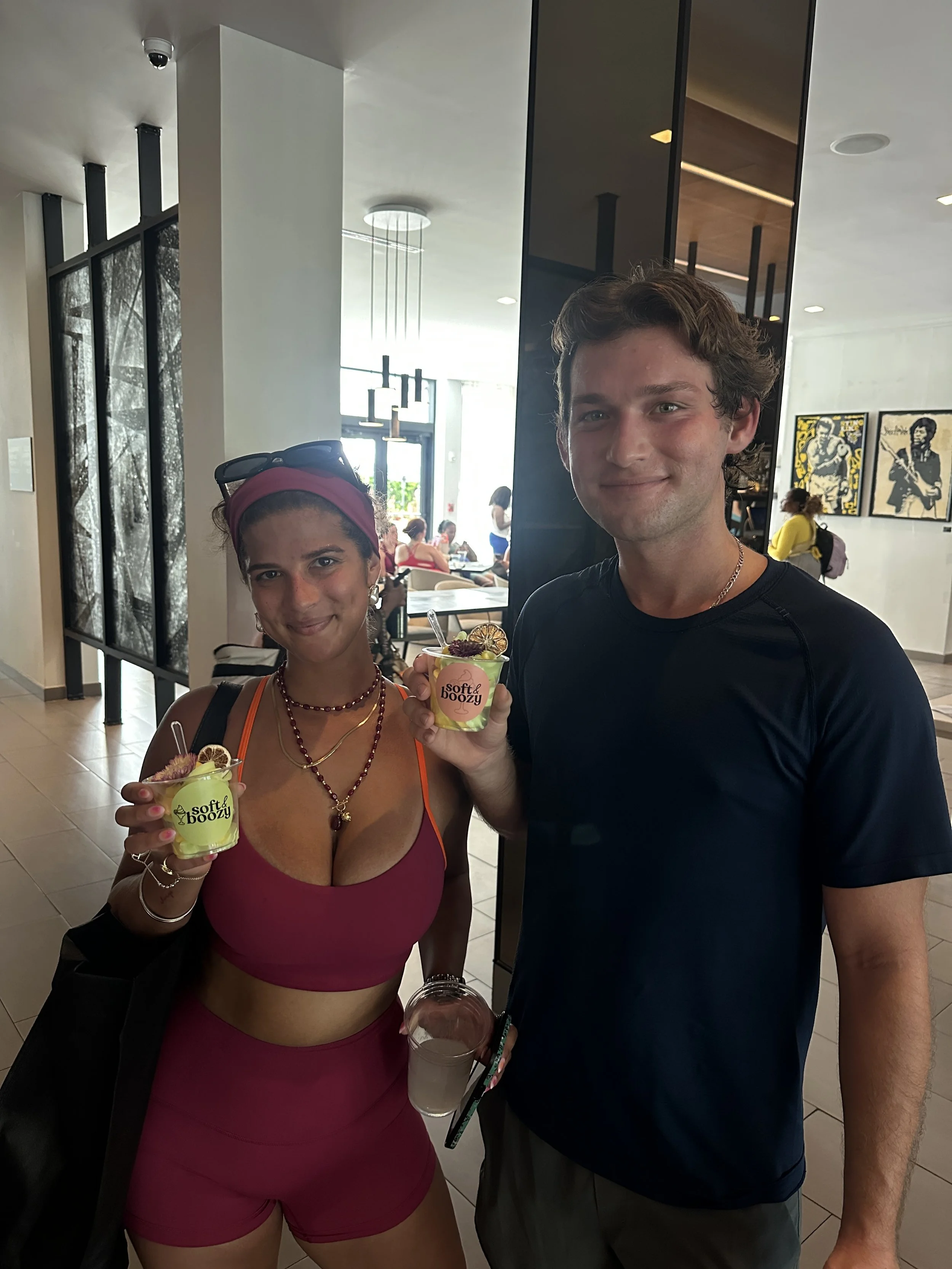 Two people holding cups of ice cream, standing inside a restaurant. The woman on the left wears a pink sports bra and shorts, has a red headband and sunglasses on her head. The man on the right wears a black t-shirt and gray shorts.