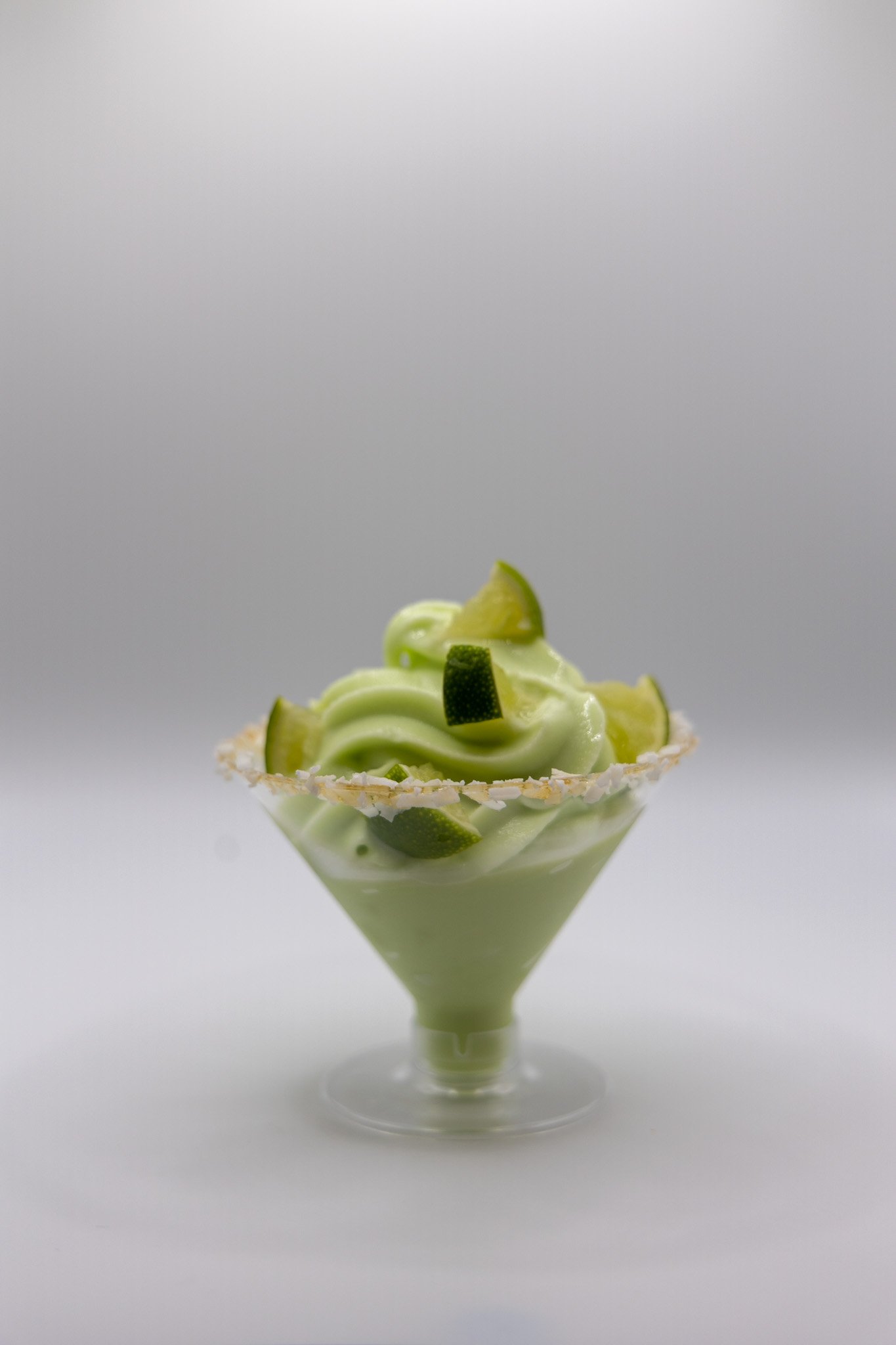 A cocktail soft-serve topped with green slices of lime inside a triangular glass bowl with a sugared rim on a white surface.