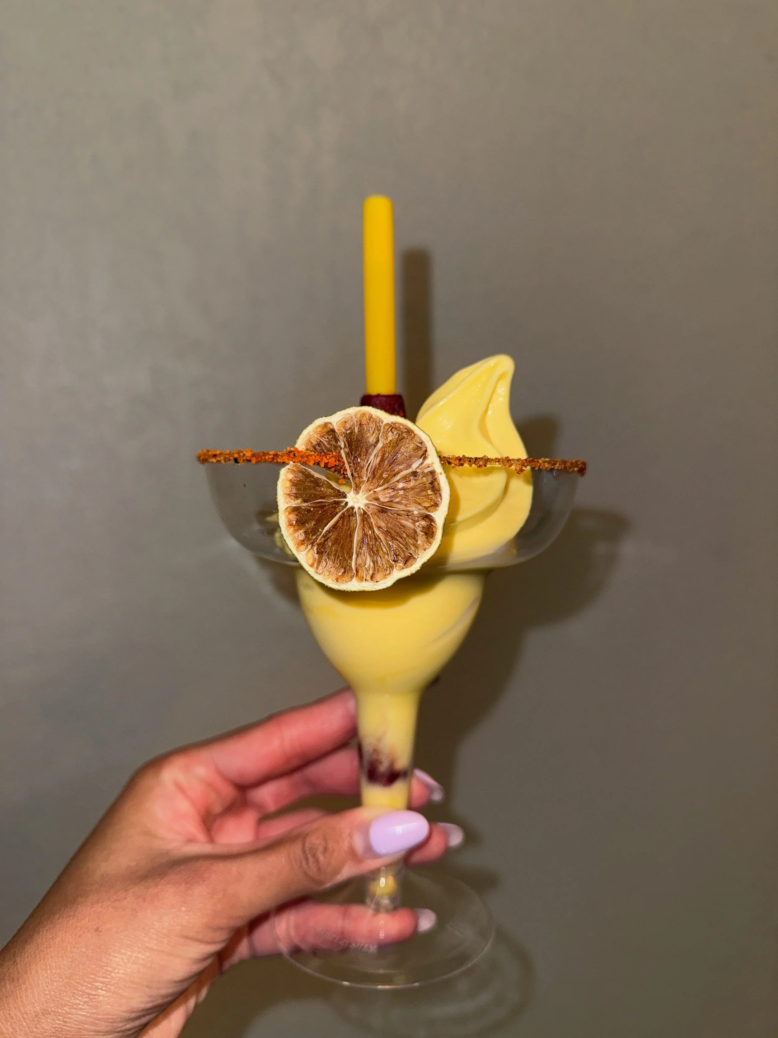 A person holding a martini glass with a yellow-colored drink, garnished with a dried lemon slice, a yellow candle, and a tajin rim decoration.