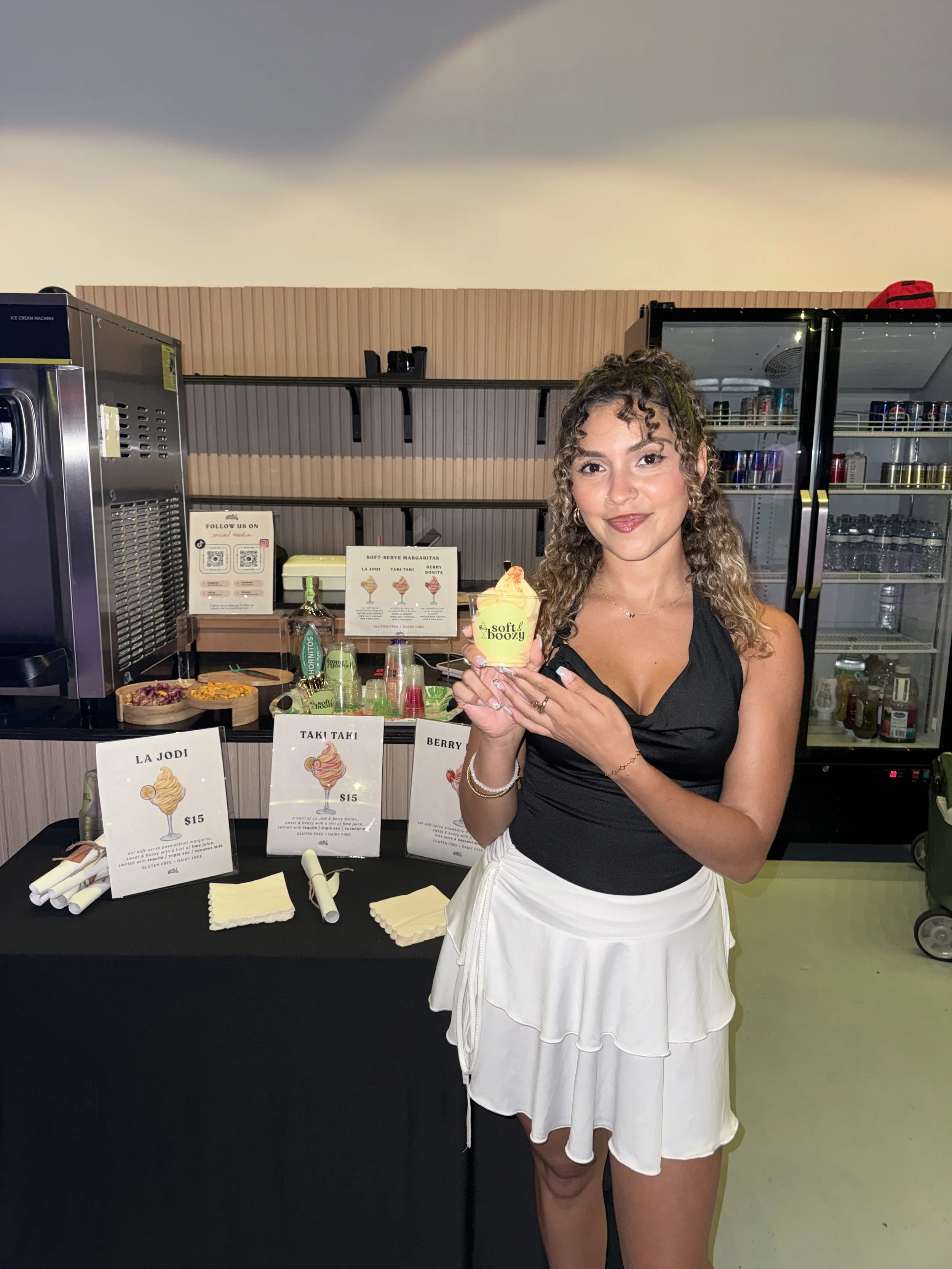 A woman with curly hair and hoop earrings, wearing a black top and white skirt, holding a yellow soft-serve ice cream in a cup labeled 'soft & boozy', standing at a food stall with menu signs for flavored soft-serve ice cream and slices of pizza.