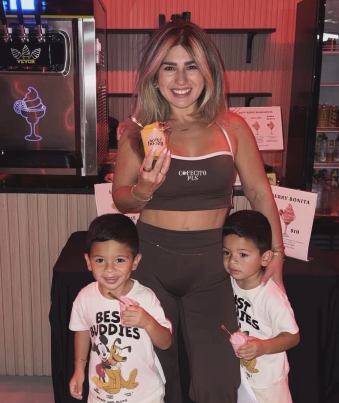 A smiling woman with shoulder-length blond hair with highlights is holding a pink ice cream cone. She is wearing a dark crop top and dark pants, standing behind two young boys with short dark hair and light brown skin. The boys are wearing white t-shirts with cartoon characters and the text "Best Buddies" and holding pink ice cream cones. They are inside an ice cream shop or stand, with signs and a menu visible in the background.