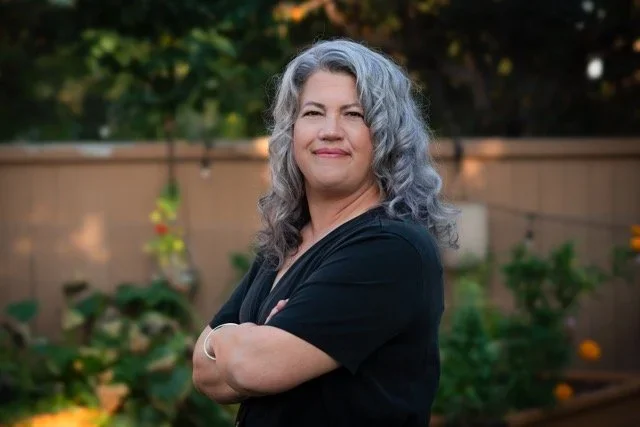 A woman with gray hair standing outdoors in front of a garden with trees and a wooden fence, wearing a black top and smiling.