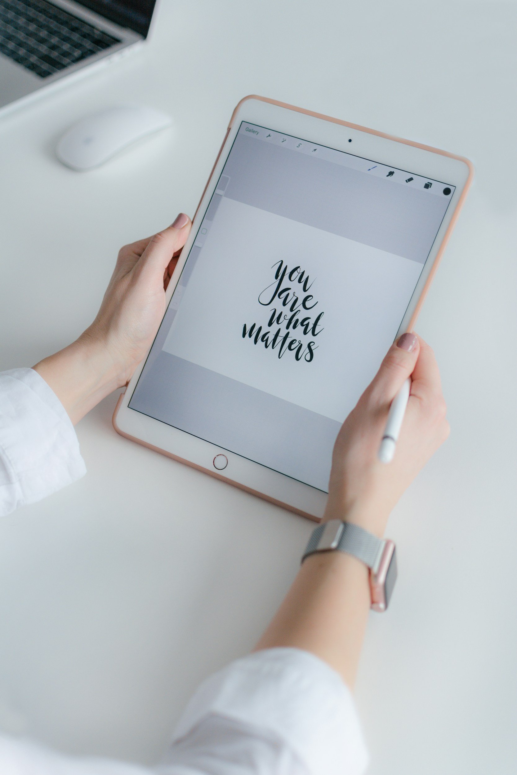 A person holding a white tablet with a motivational quote displayed on the screen that says "you are what matters" in black cursive font. The person is wearing a grey watch and a white long-sleeve shirt, and the tablet is on a white table with a laptop and wireless mouse nearby.