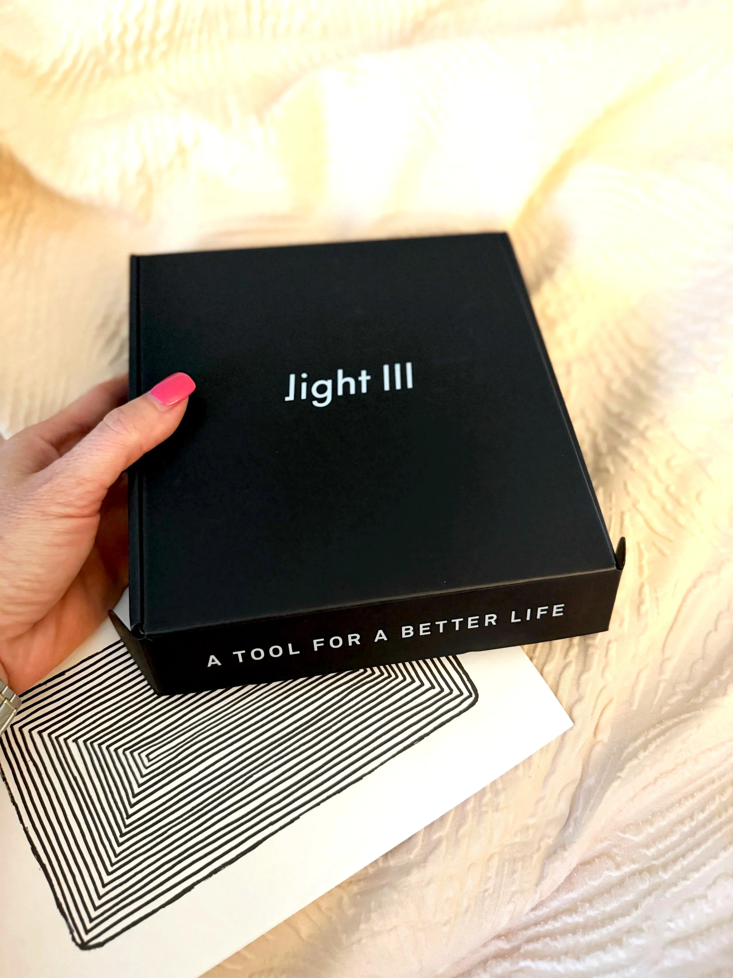 A person with pink nail polish holding a black box labeled 'Light III.' The box is on a table with a white paper and a black-and-white patterned zine.