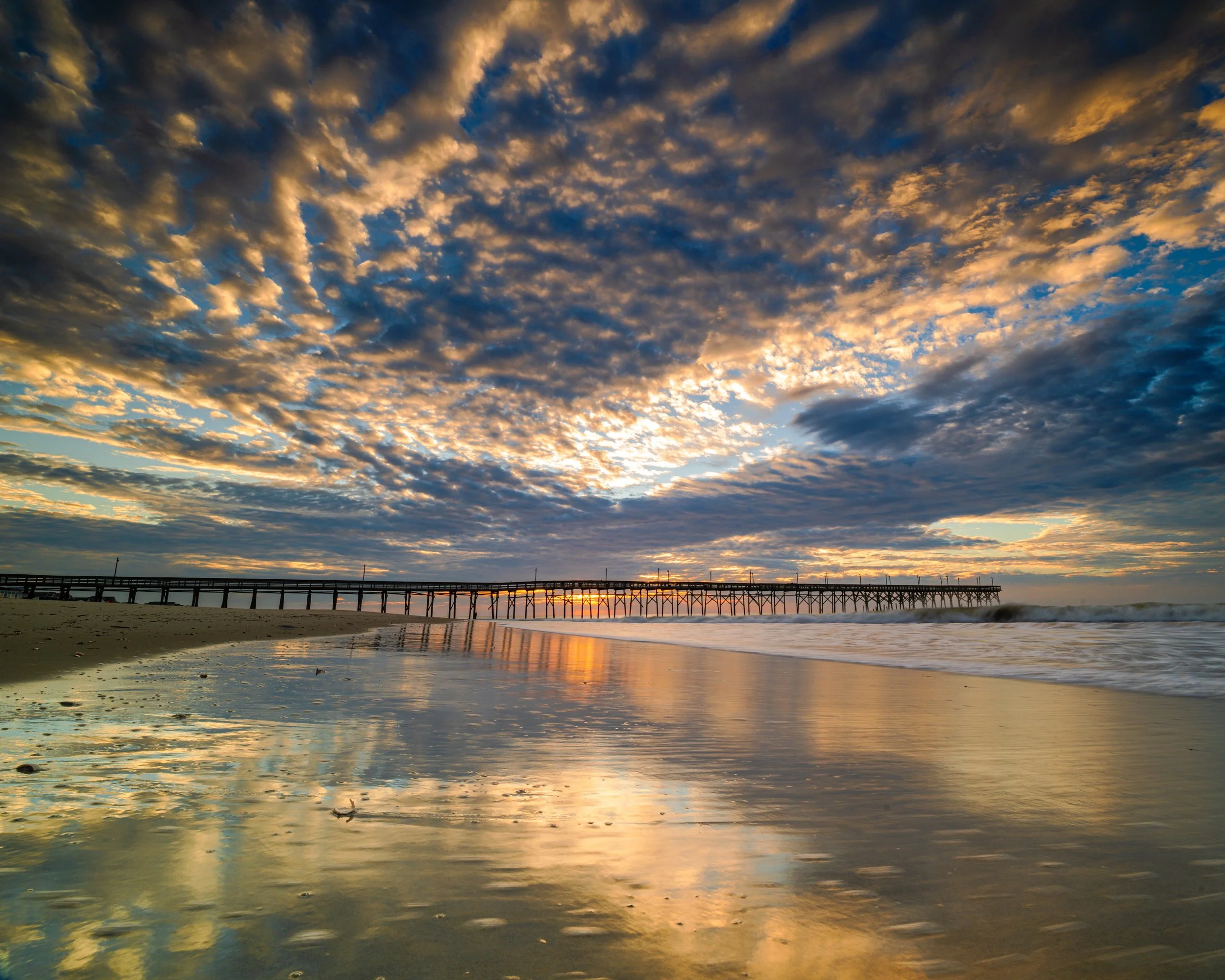 Sunrise reflecting across wet sand beneath Oak Island Pier in Brunswick County, North Carolina, as dramatic morning clouds catch the first light of day.