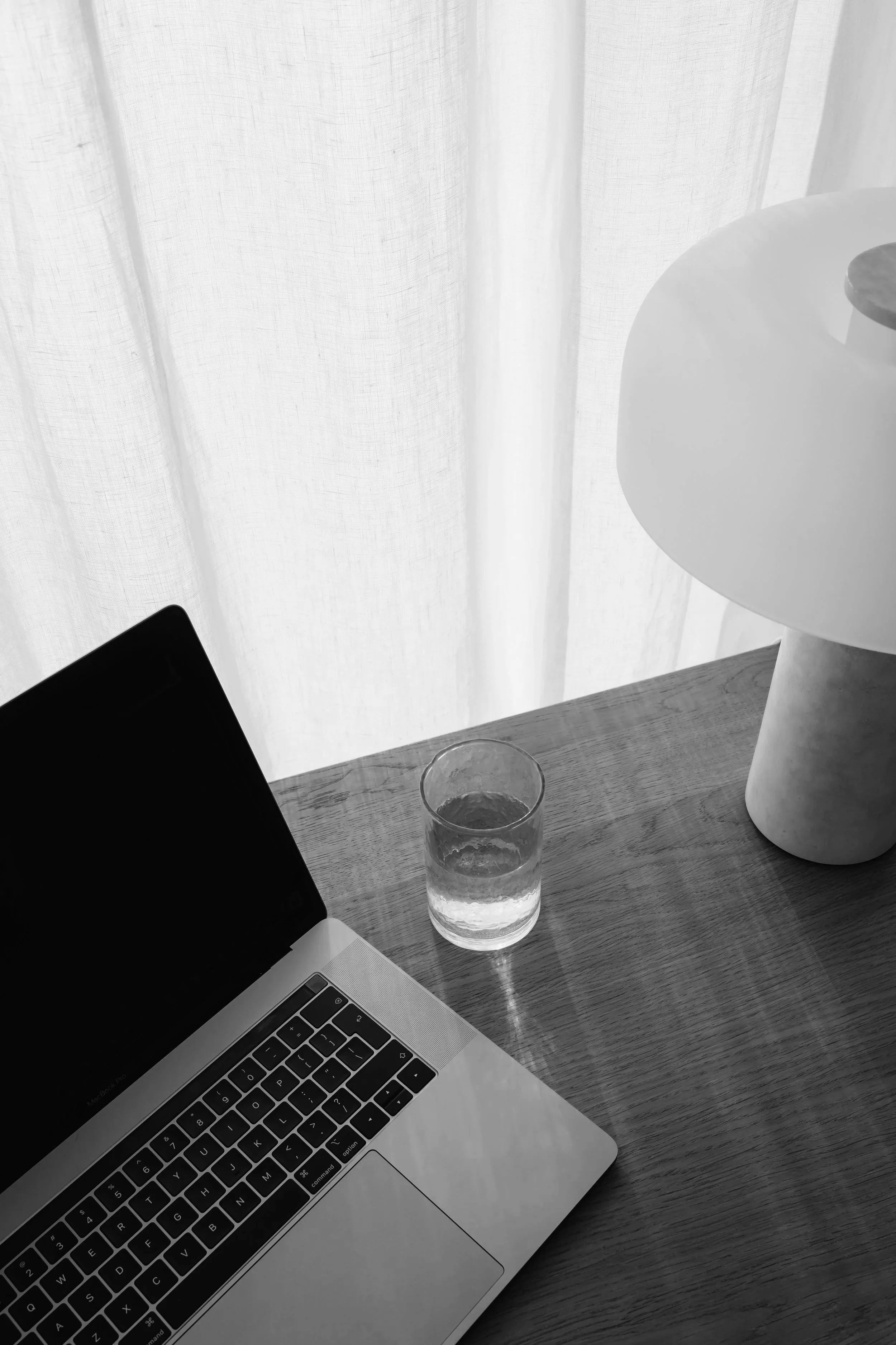 A workspace with a laptop, a glass of water, and a unique white table lamp on a wooden desk. The background has sheer white curtains.