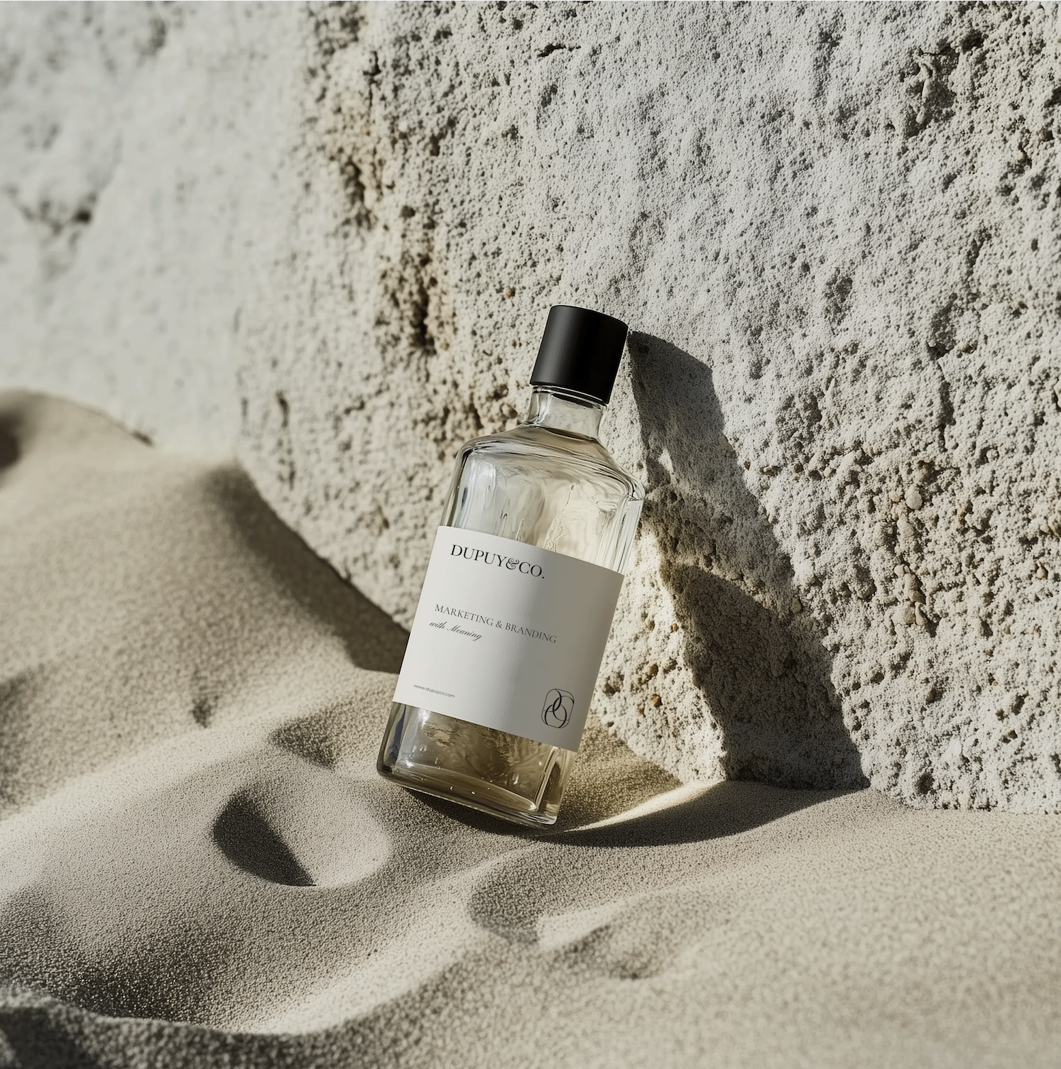 A clear glass bottle of perfume with a black cap, labeled 'DUPUY & CO.' and 'MARKETING & BRANDING with Mearing,' lying on sandy ground with a large textured rock in the background, casting a shadow.