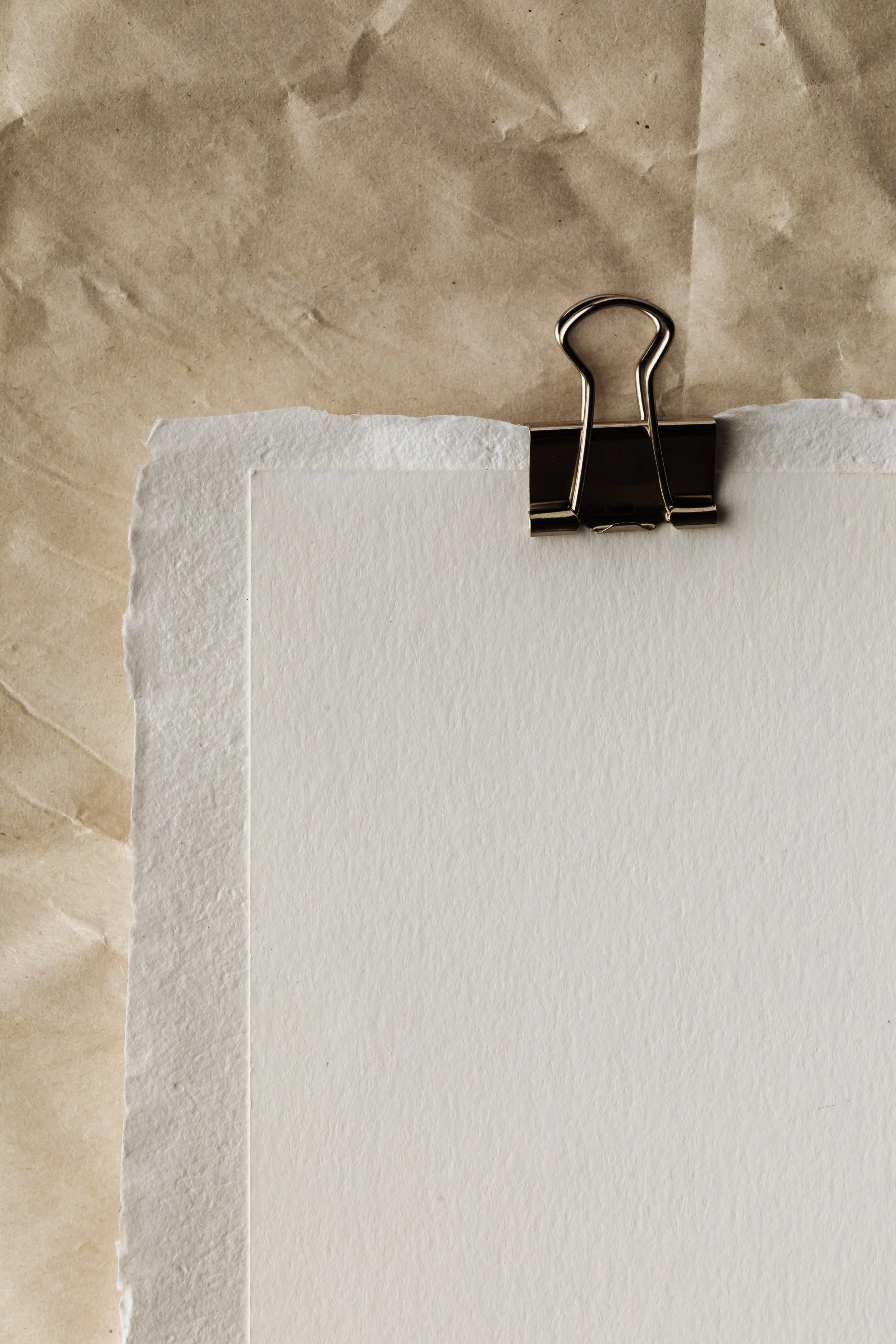 Close-up of a binder clip holding a piece of white textured paper on top of a brown paper background.