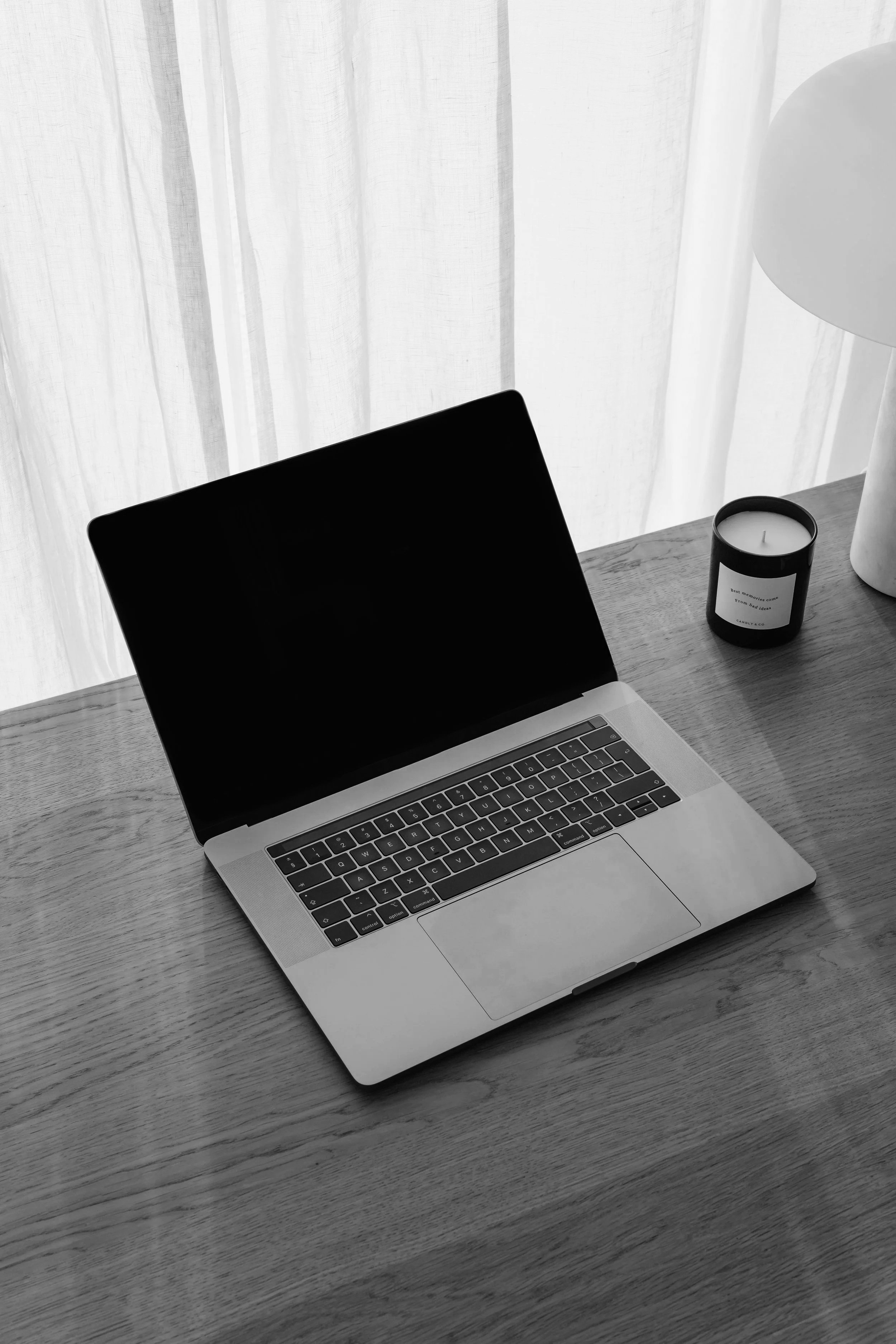 A silver laptop with a black screen on a wooden desk, next to a black candle with a white label and a white table lamp in front of a sheer white curtain.