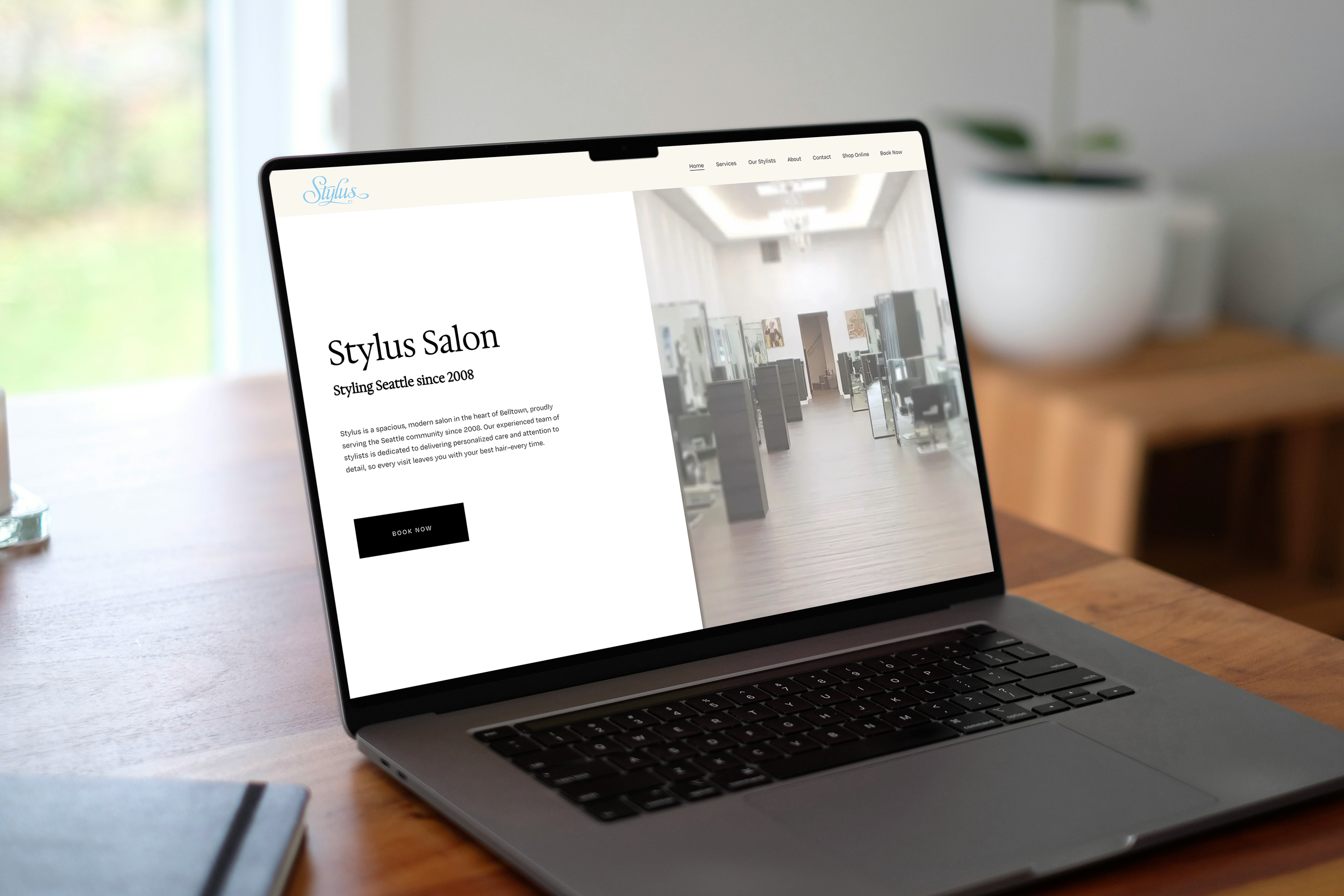 A laptop on a wooden table displaying a website for Stylus Salon with a hair salon interior image.