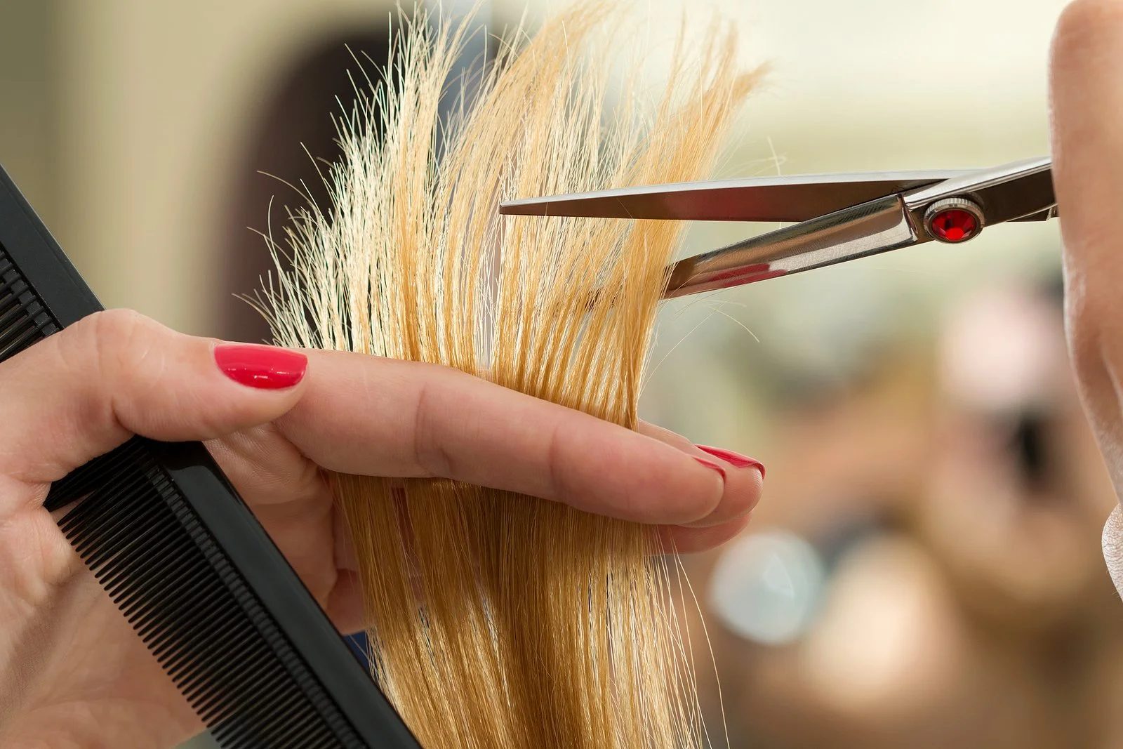A hairdresser trims a person's blond hair with scissors, holding a section of hair between fingers and using a black comb to section it.