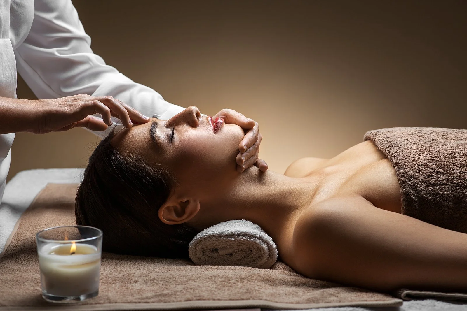 A woman is getting a facial massage at a spa, lying on a massage table with her eyes closed, with a towel across her chest. A therapist is gently massaging her face with both hands. A lit candle is in the foreground, contributing to a relaxing ambiance.