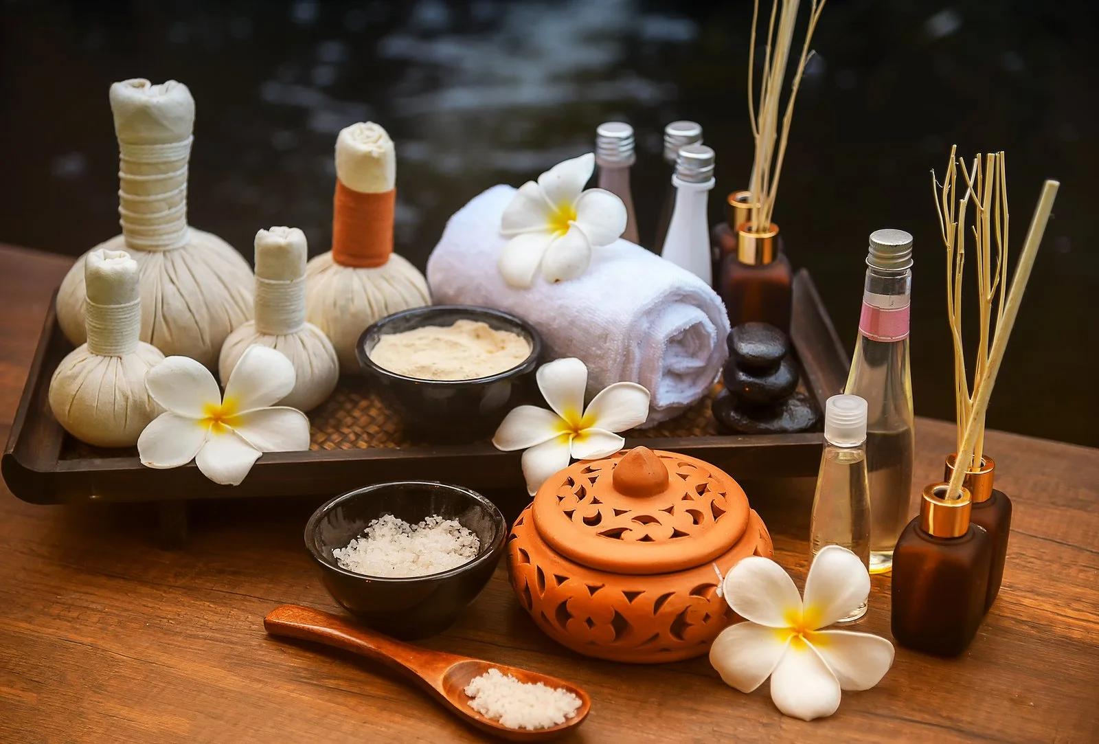Spa treatment setup with herbal compress balls, rolled towels, flowers, massage oils, bath salts, and spa accessories on a wooden surface.