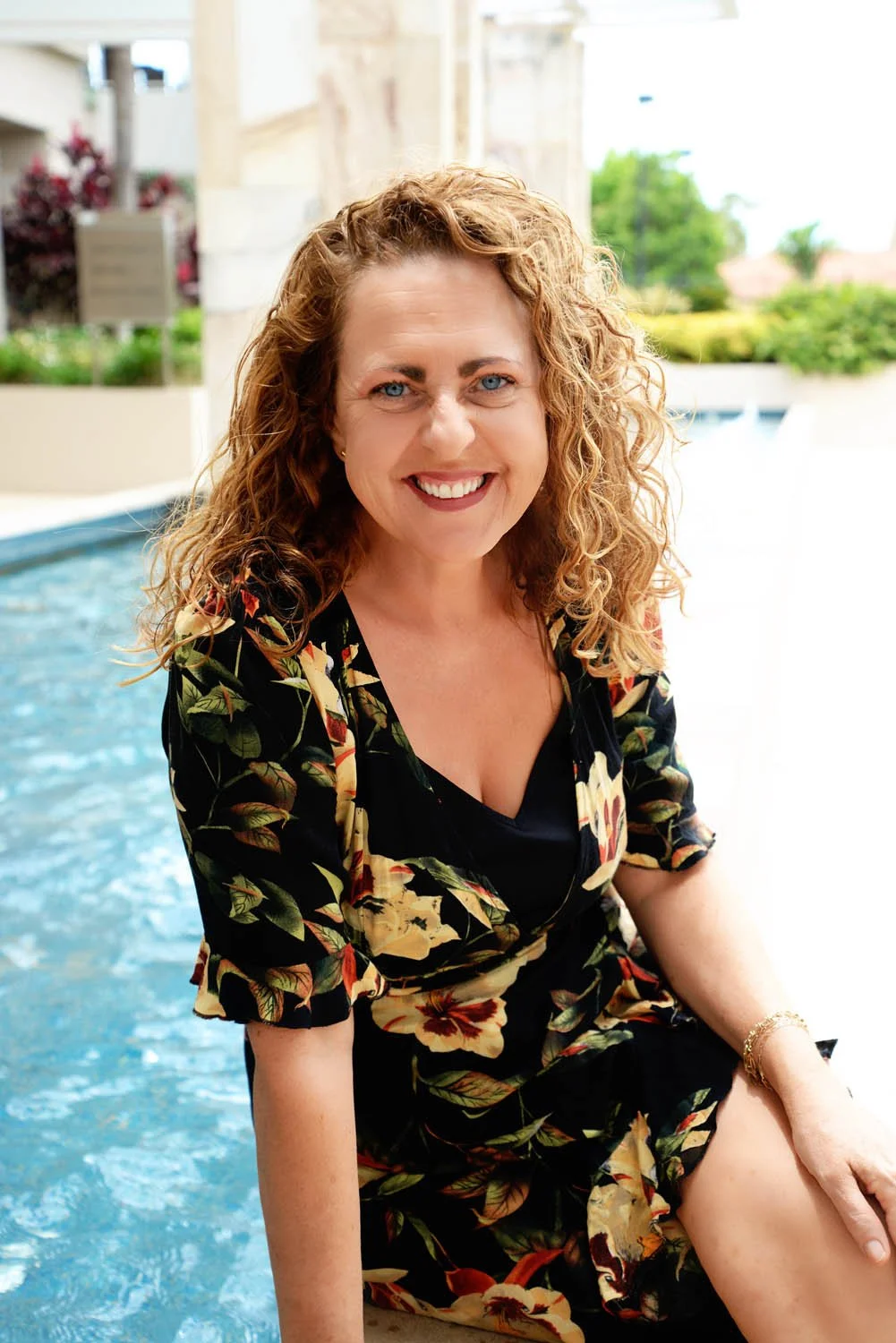A woman with curly red hair and blue eyes smiling at the camera, sitting by a pool on a sunny day, wearing a black floral dress.