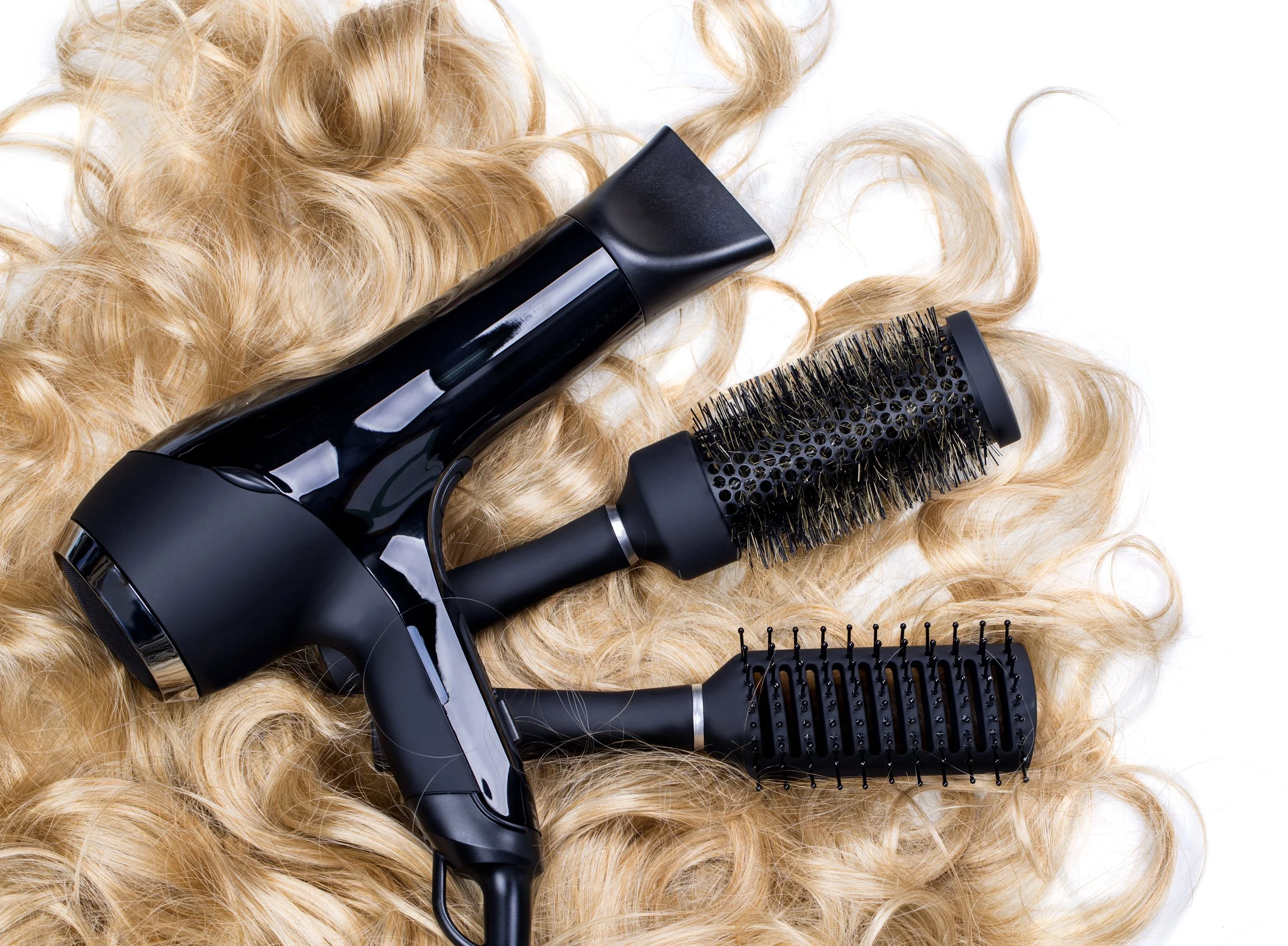 A black hairdryer with a shiny surface, a round brush, and a black vented hairbrush are placed on long, curly blonde hair.