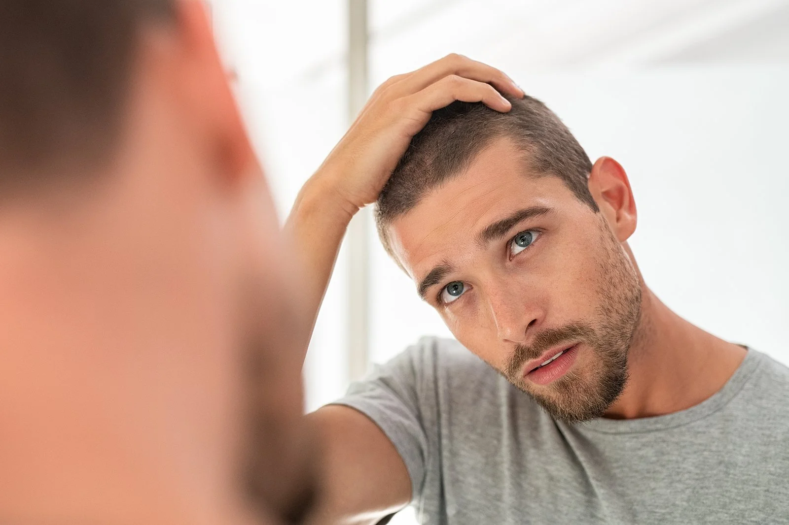 A young man with short dark hair and a beard looking in the mirror, touching his head with his right hand, in a well-lit room.