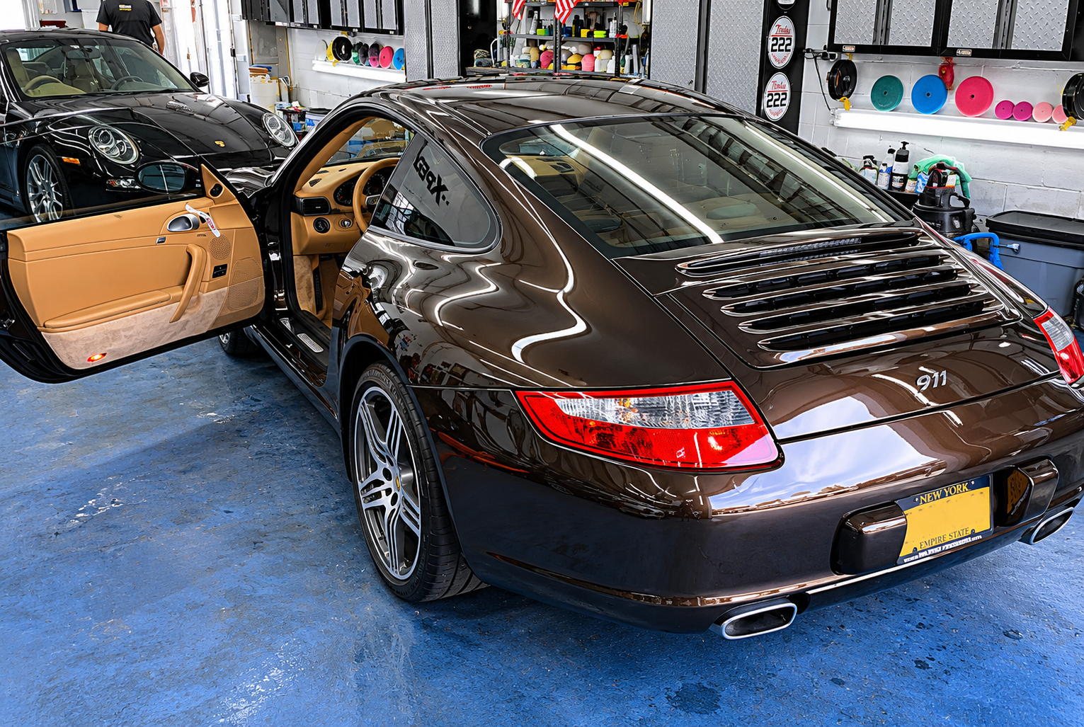 A black Porsche 911 with a tan interior parked inside a garage, with the driver's side door open, revealing the interior. Another black Porsche is partially visible in the background.
