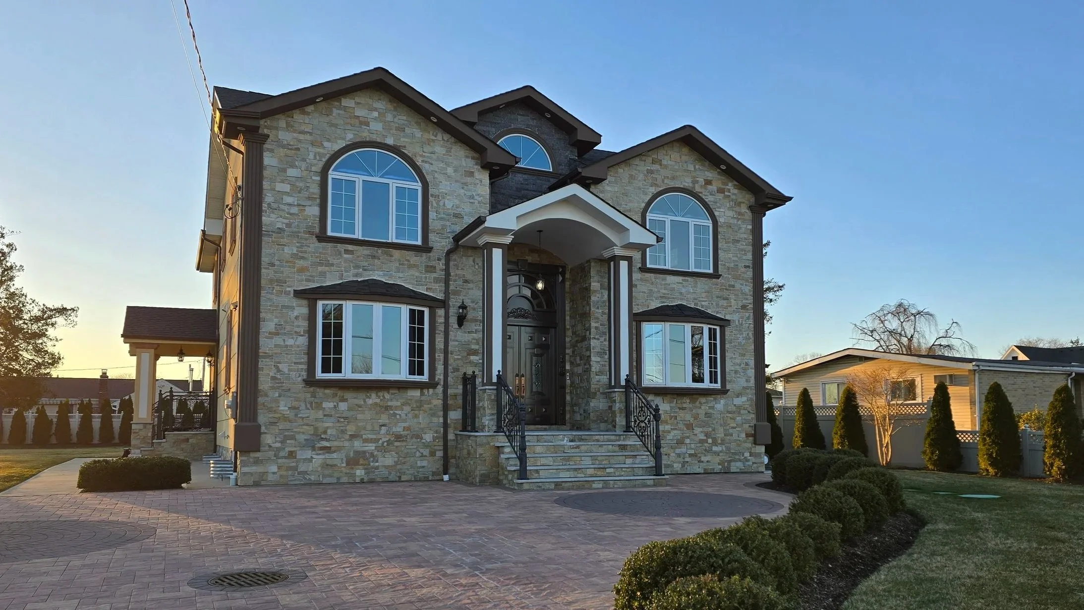 A large, multi-story house built with stone and siding, featuring arched and rectangular windows, a front staircase, and a landscaped yard with bushes and trees.