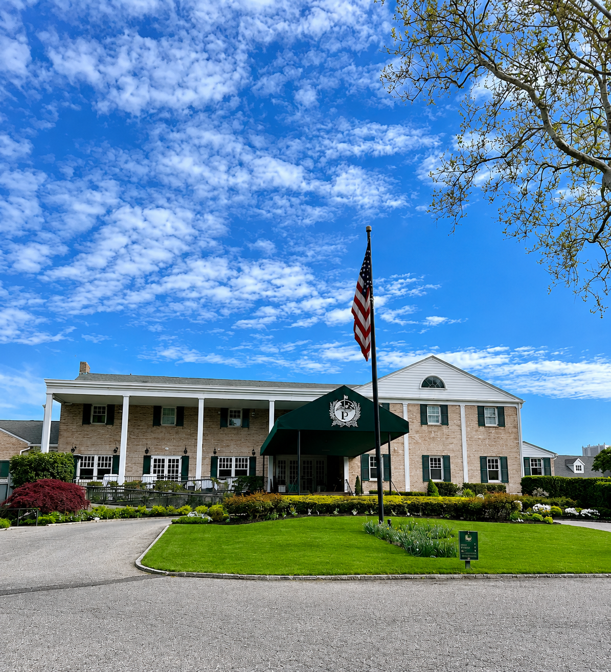 A two-story brick building with a white triangular roof, green shutters, and a covered entrance with a green awning featuring a white emblem. An American flag on a flagpole is in front, surrounded by a well-manicured lawn and garden. Blue sky with scattered clouds overhead.