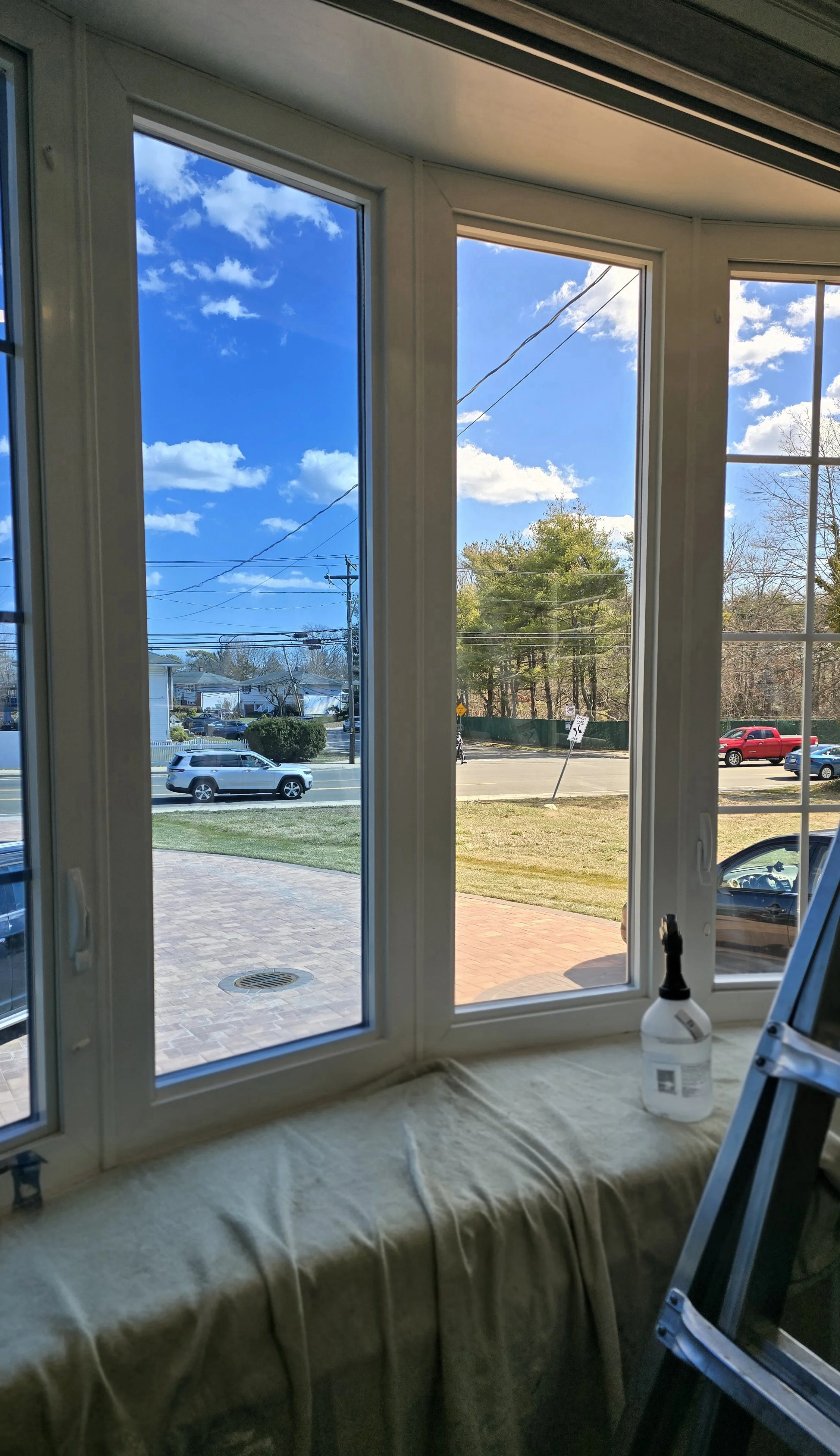 Looking outside a large window, a quiet suburban street with cars parked and driving by, trees with green leaves, power lines, a sidewalk, and a blue sky with some clouds.