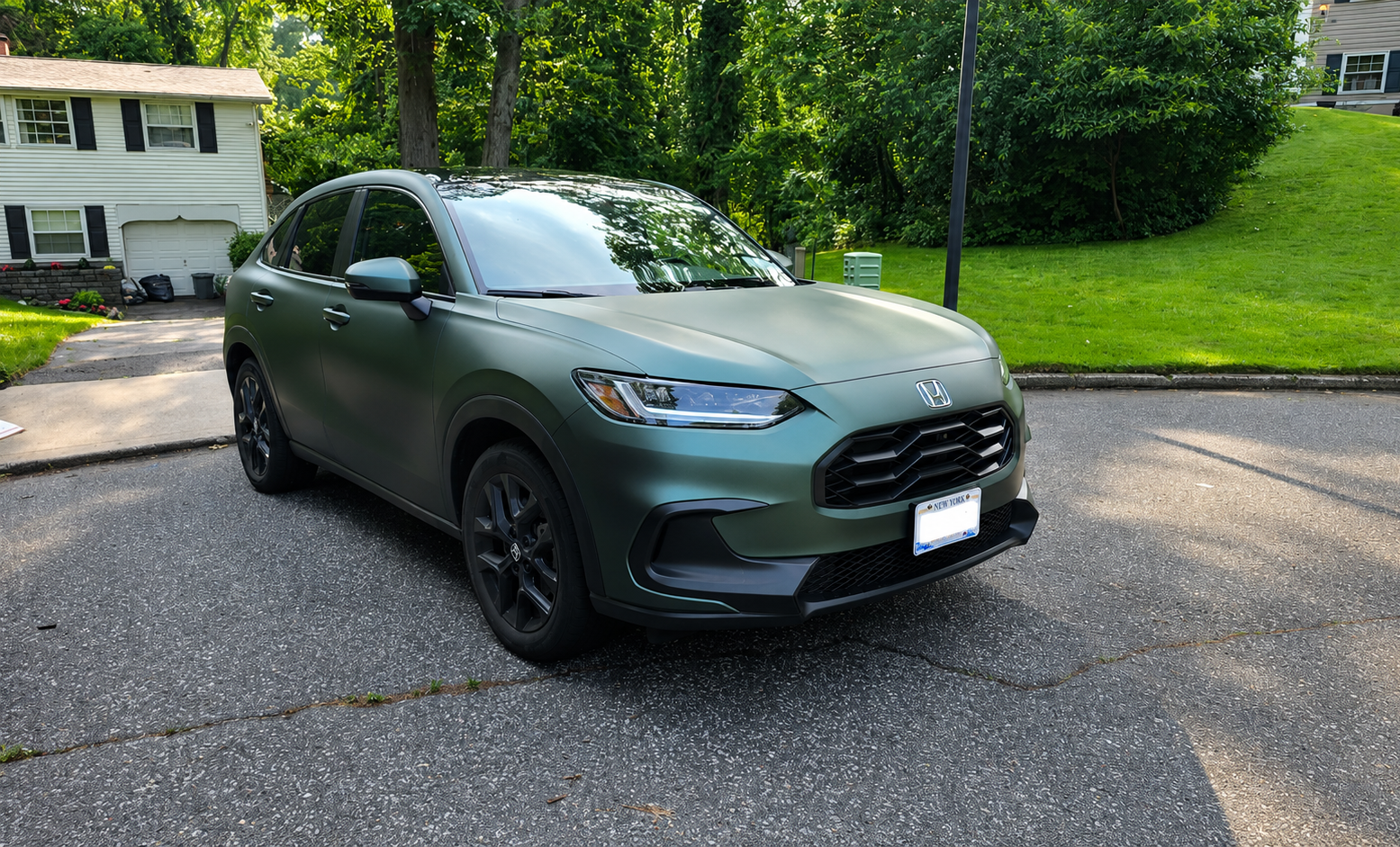 A matte green Honda SUV parked on a residential driveway with green grass, trees, and houses in the background.