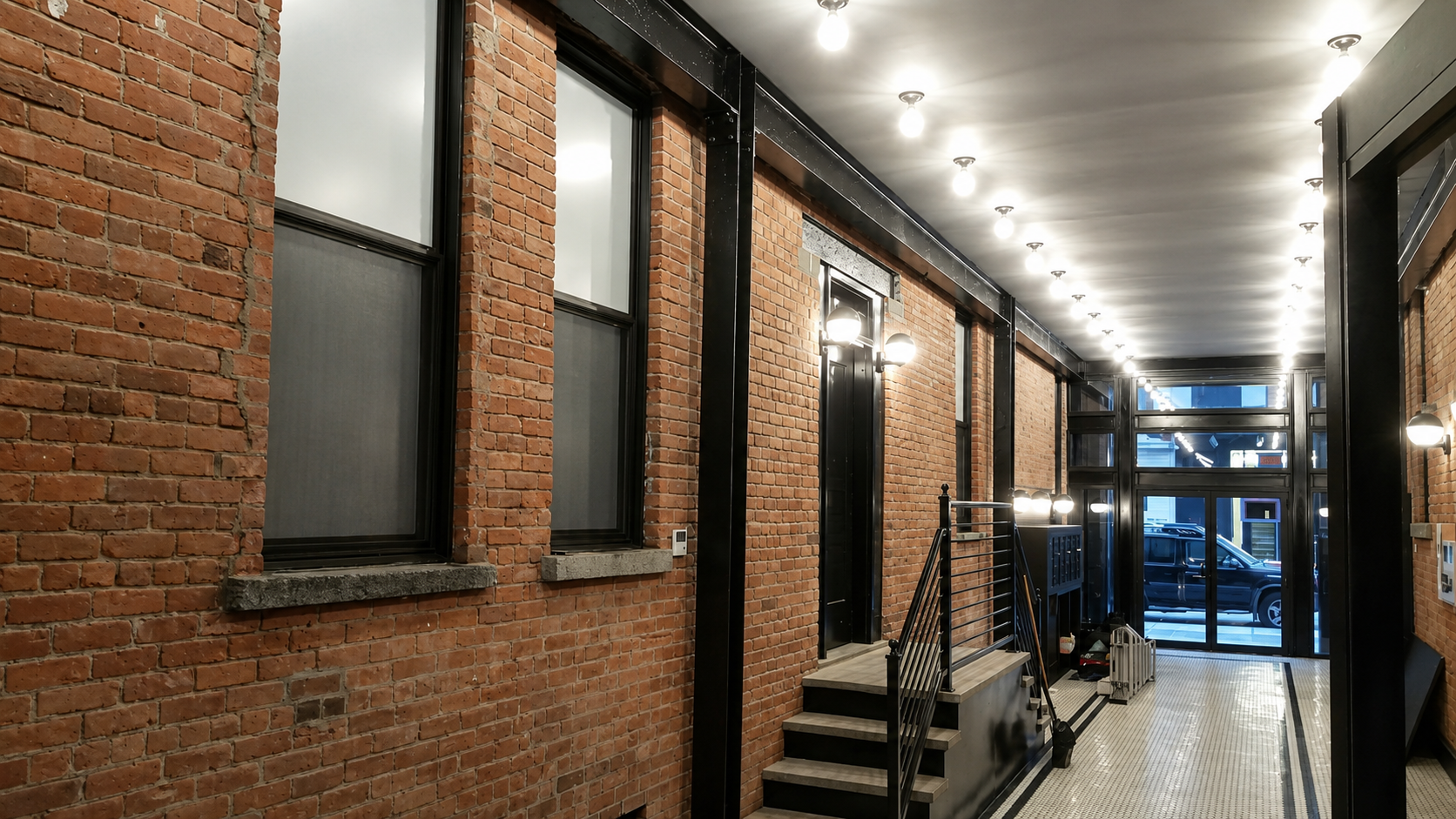 Indoor hallway with exposed red brick walls, black window frames, and metal accents, illuminated by ceiling bulbs and wall-mounted lights, leading to a glass door showing a parked vehicle outside.
