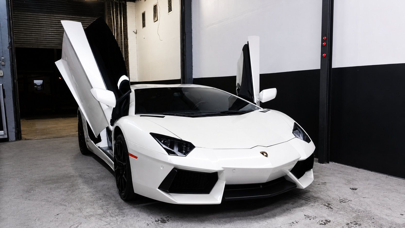A white Lamborghini supercar with scissor doors open, parked indoors in a garage or showroom.