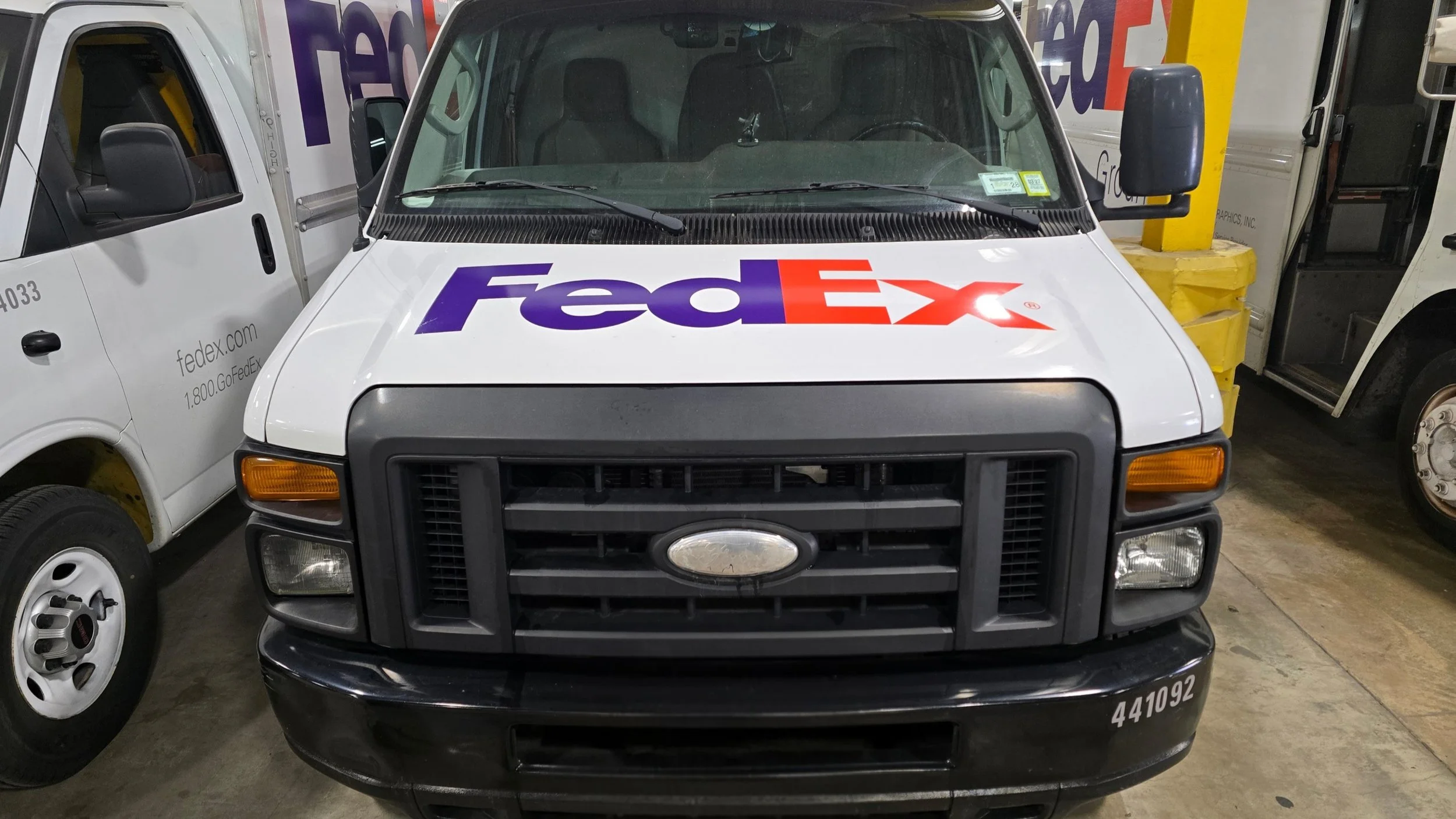 Front view of a FedEx delivery vehicle parked inside a building, flanked by other white delivery vans.