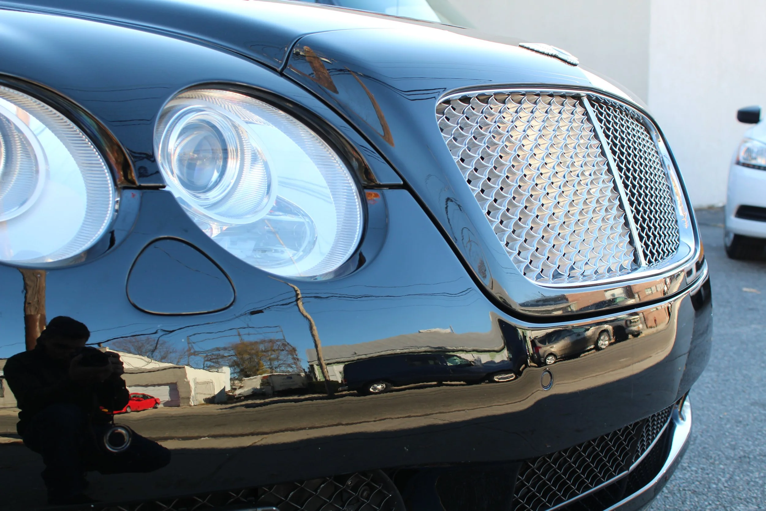 Close-up of the front of a black luxury car, showing the headlights, grille, and part of the bumper. The car's shiny surface reflects nearby objects and the sky.