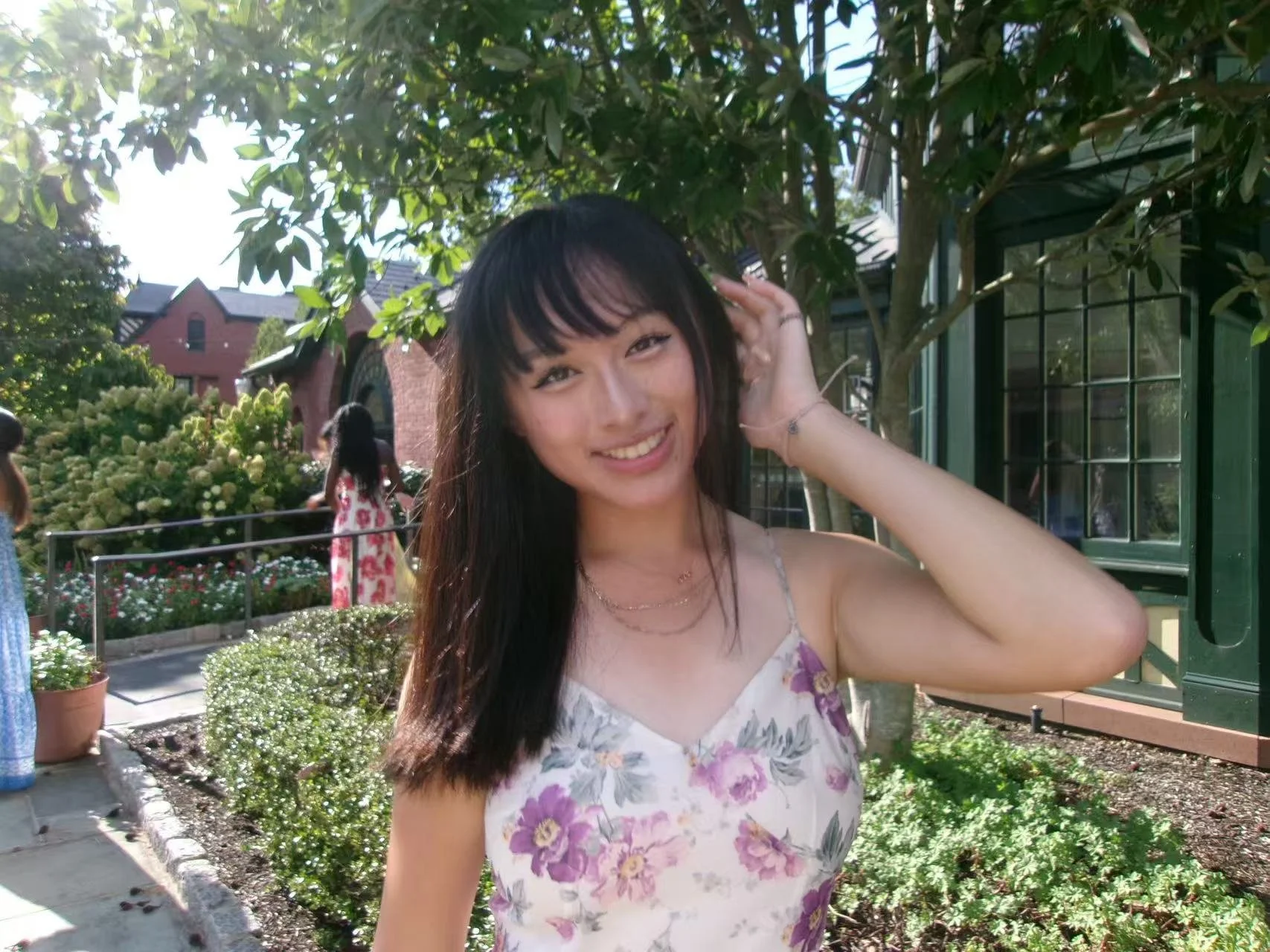 Smiling young woman with long dark hair, wearing a floral dress, posing outdoors in a garden area with trees, flowers, and a green building in the background.