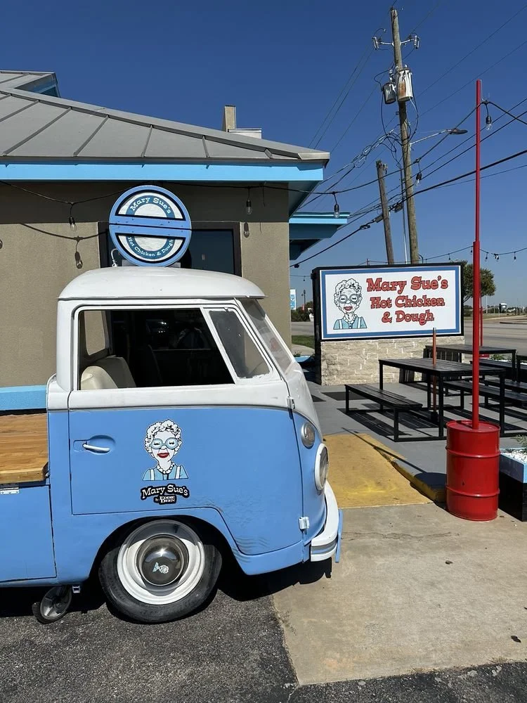 A blue and white vintage auto rickshaw with a Mary Sue's Hot Chicken & Dough sign and logo parked outside a restaurant with a sign and outdoor seating area.