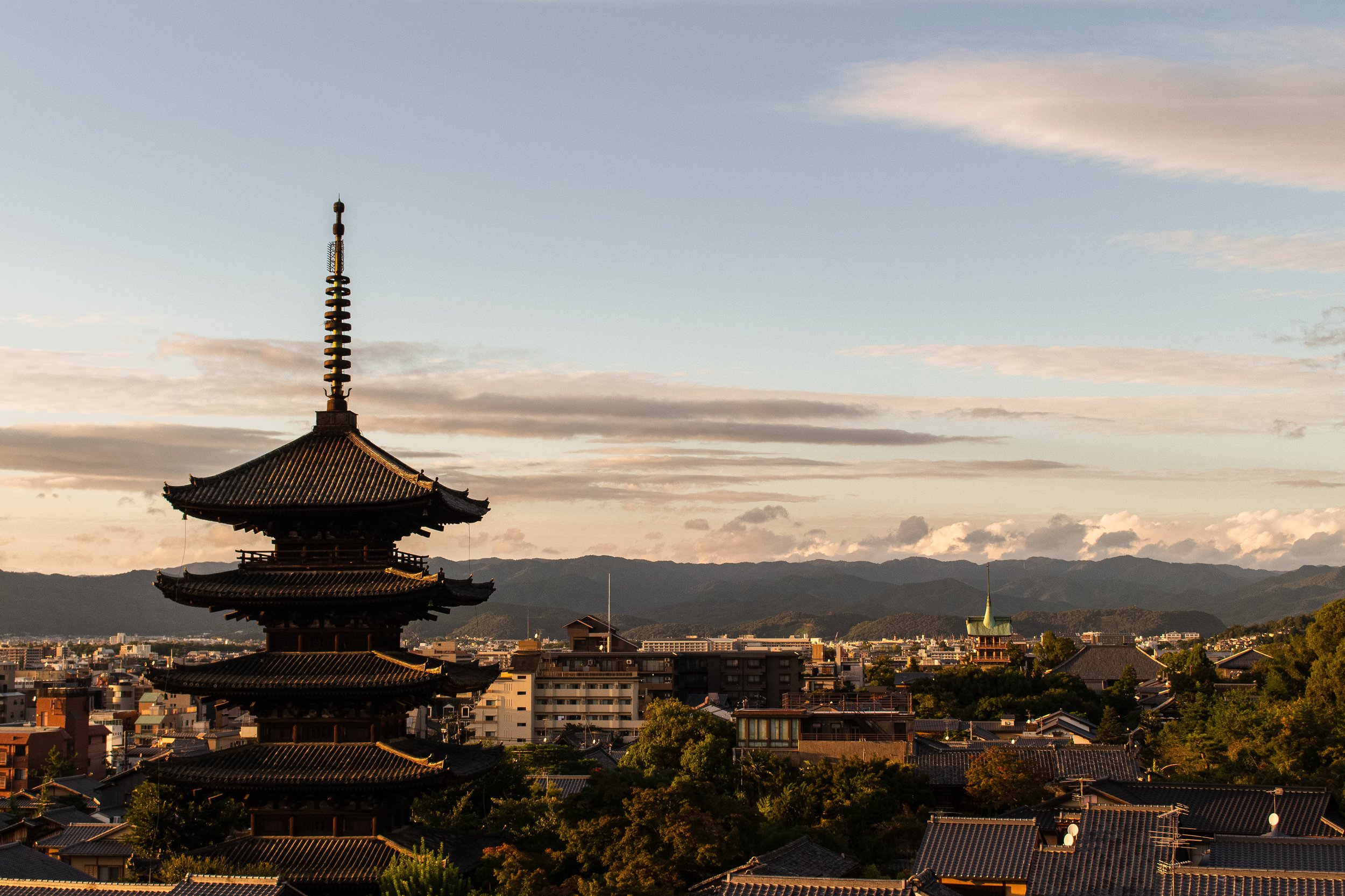 A traditional Japanese pagoda with a multi-tiered roof and a tall spire, set against a backdrop of mountains and a partly cloudy sky during sunset.