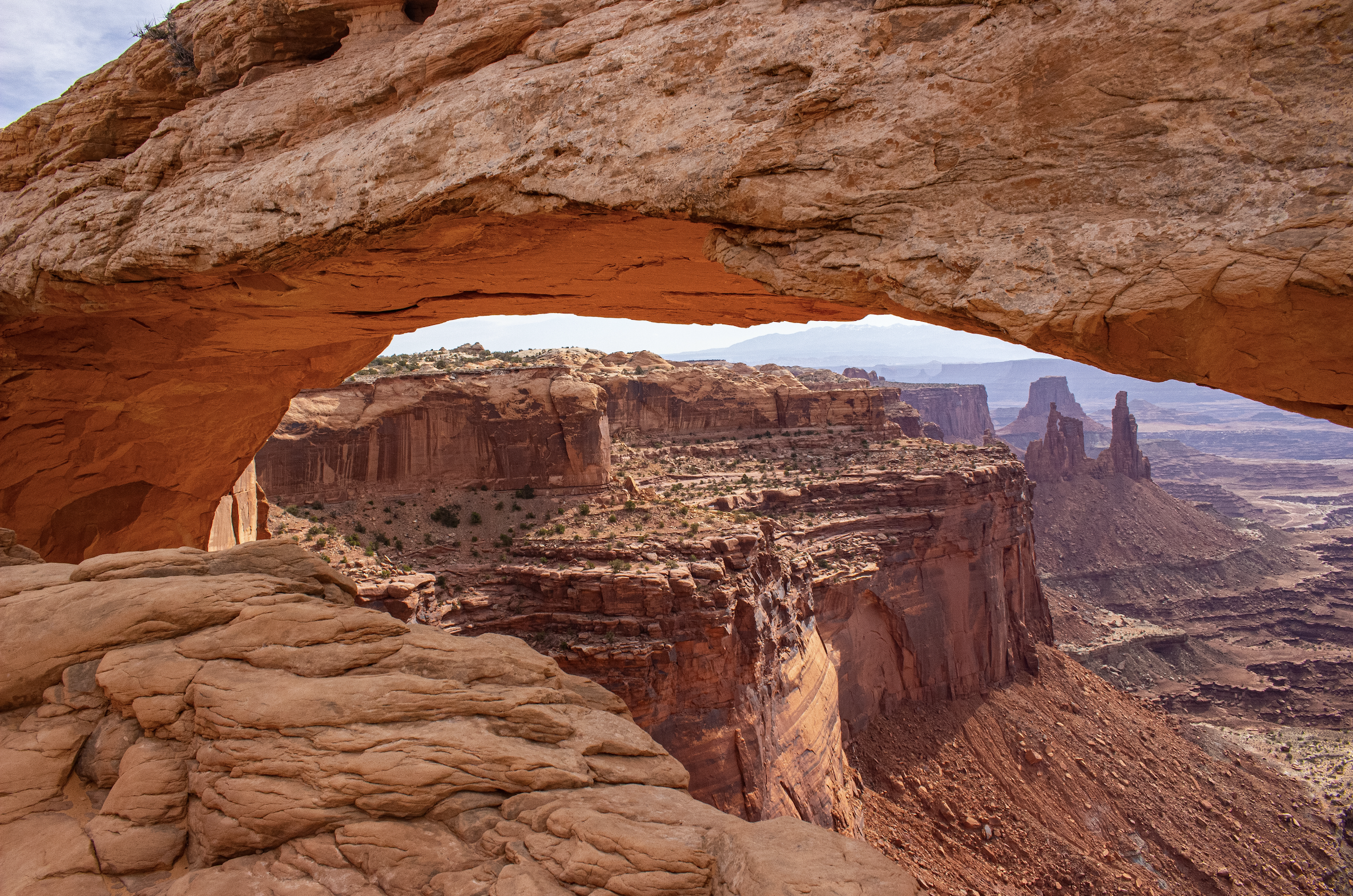 Natural rock arch in a desert landscape with rugged red canyon walls and distant plateaus under a partly cloudy sky.
