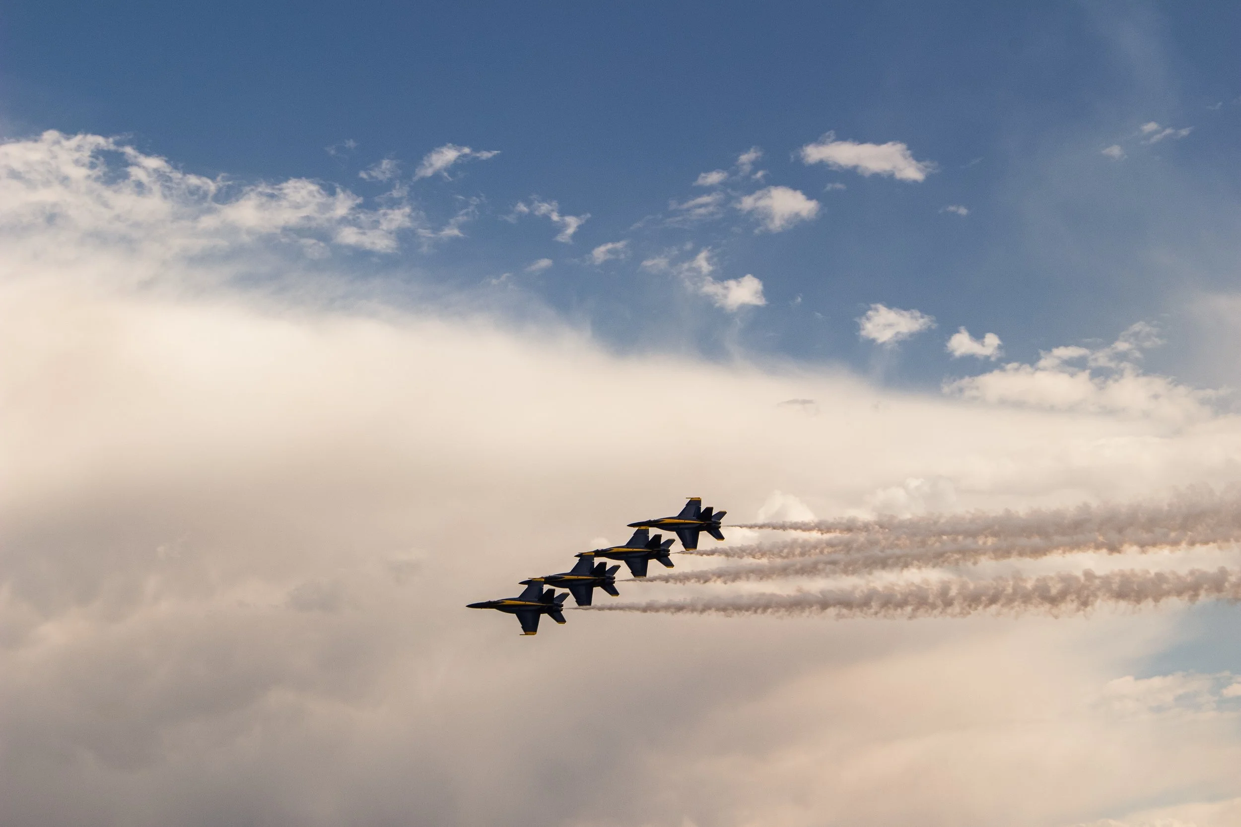 A formation of jet fighters flying in a diagonal line across the sky, leaving smoke trails behind them, with clouds and blue sky in the background.