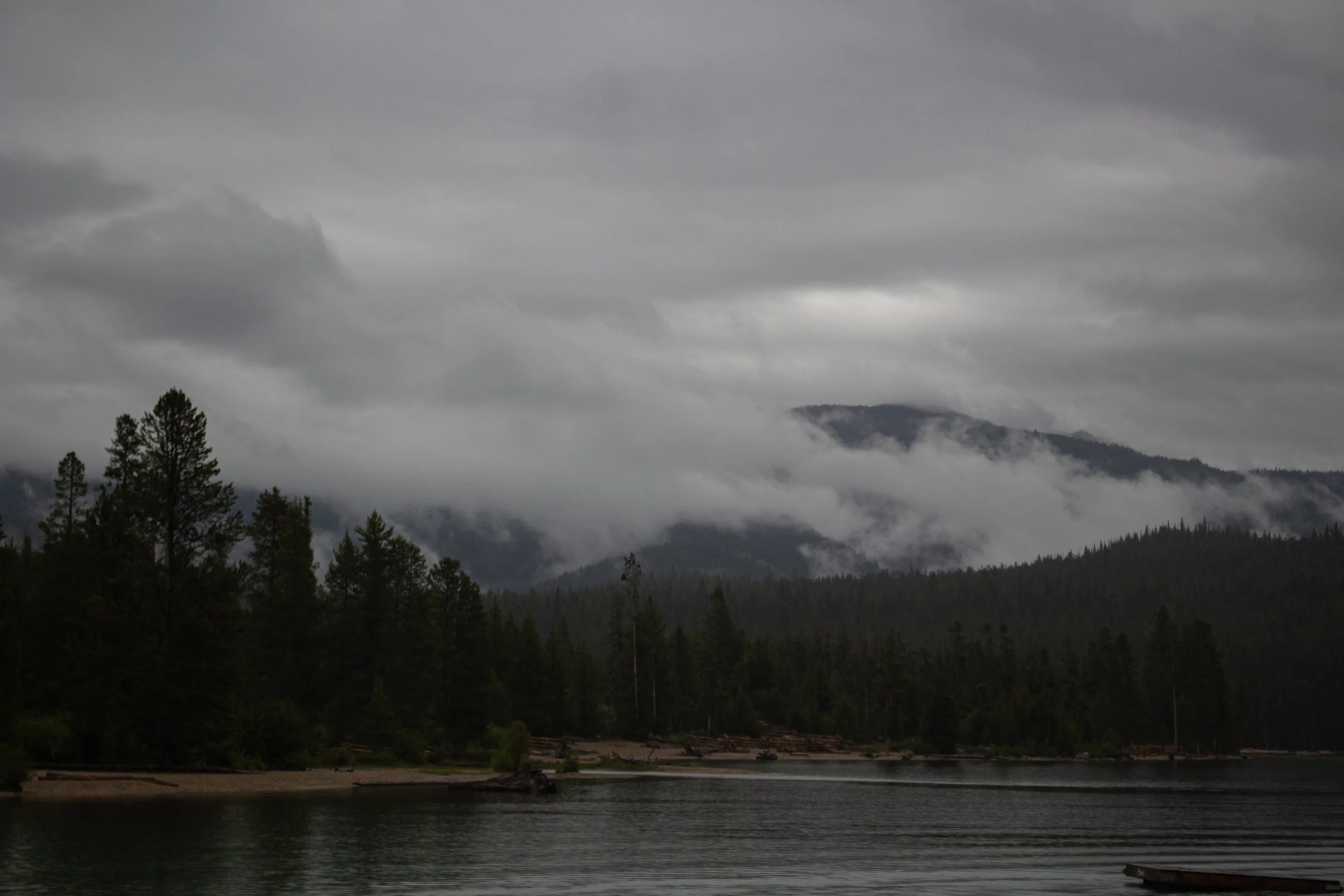 Overcast sky with dark clouds over a forested mountain landscape and a body of water in the foreground.
