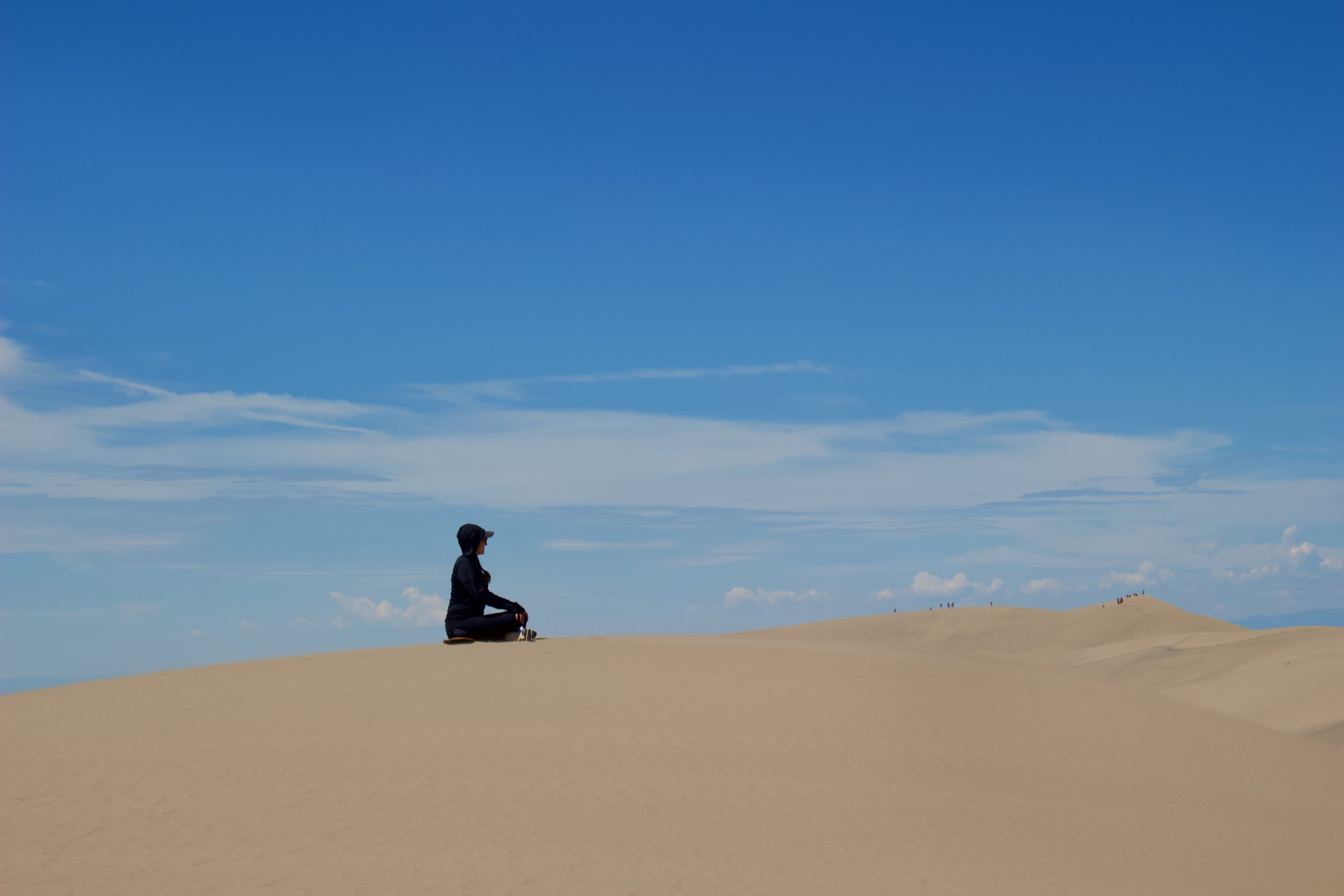 A person sitting cross-legged on sand dunes, looking toward the horizon under a blue sky with few clouds, with small groups of people visible in the distance.