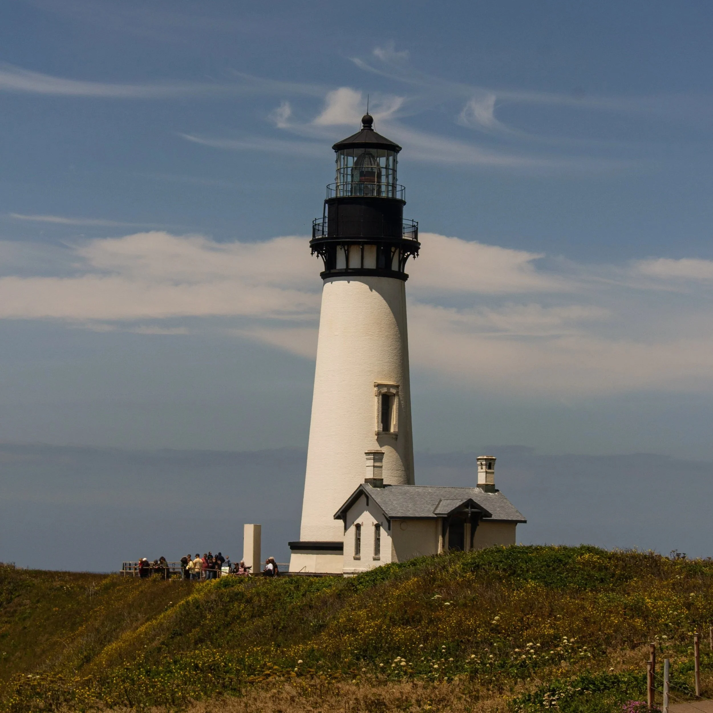 Lighthouse on a grassy hill with a group of people nearby against a partly cloudy sky.