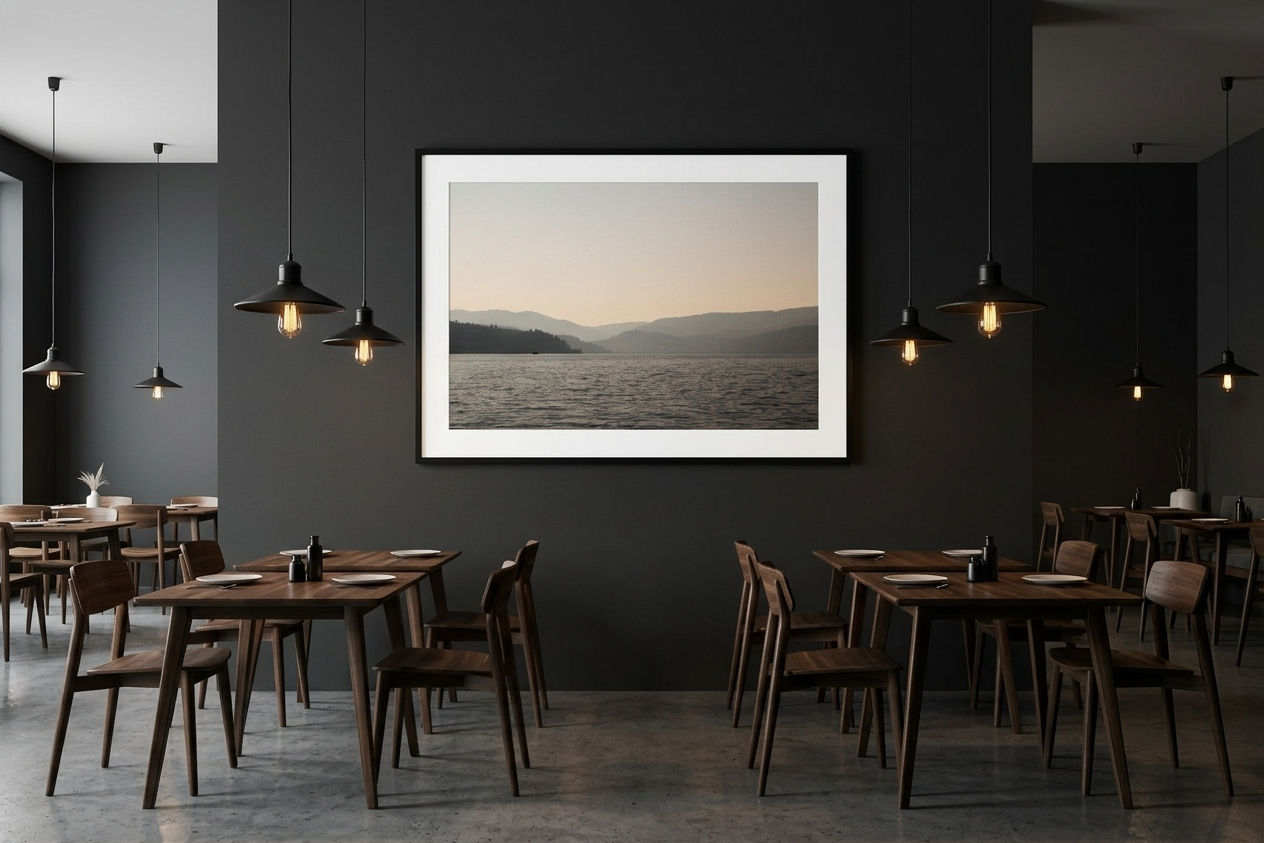 A modern restaurant interior with dark gray walls, wooden tables and chairs, pendant lighting, and a large framed landscape photograph of a lake and mountains on the wall.