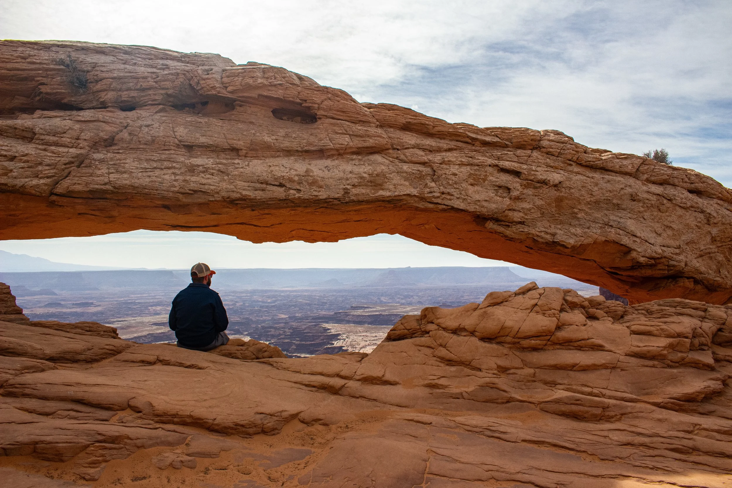 A person with a cap and jacket sitting on a rock formation under a large natural arch in a desert landscape with distant mountains and cloudy sky.