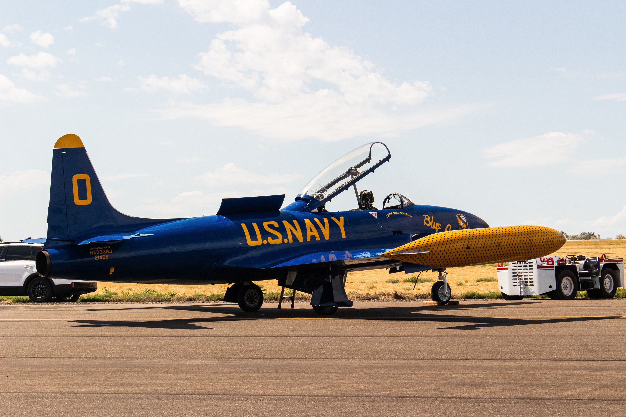 A blue US Navy fighter jet parked on the tarmac with a yellow and black missile under its wing. The plane's cockpit canopy is open, and there are vehicles and equipment nearby. The sky is blue with some clouds.