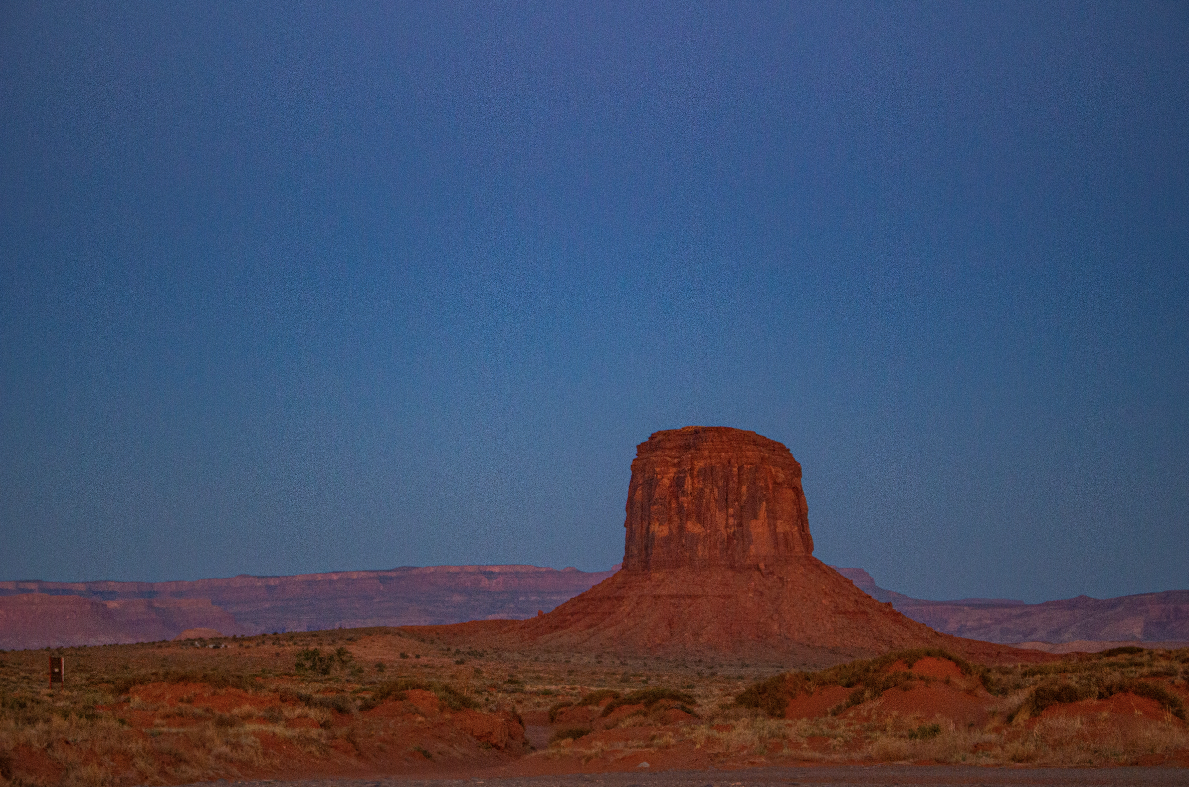 A tall, red rock formation in a desert landscape during twilight with a clear blue sky and distant mountains.