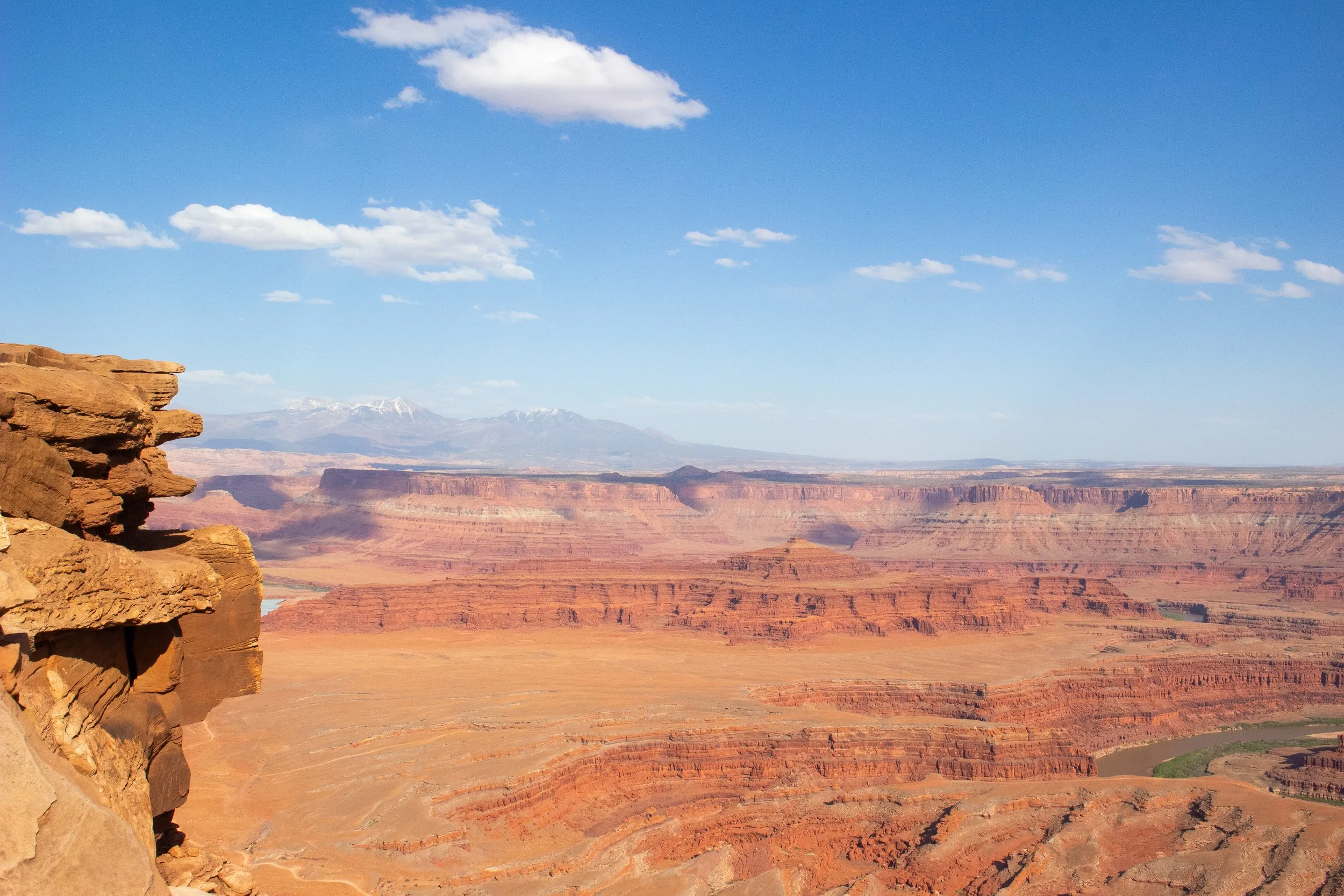 View of the Grand Canyon with layered red rock formations, distant snow-capped mountains, a cloudy blue sky, and a rocky ledge in the foreground.