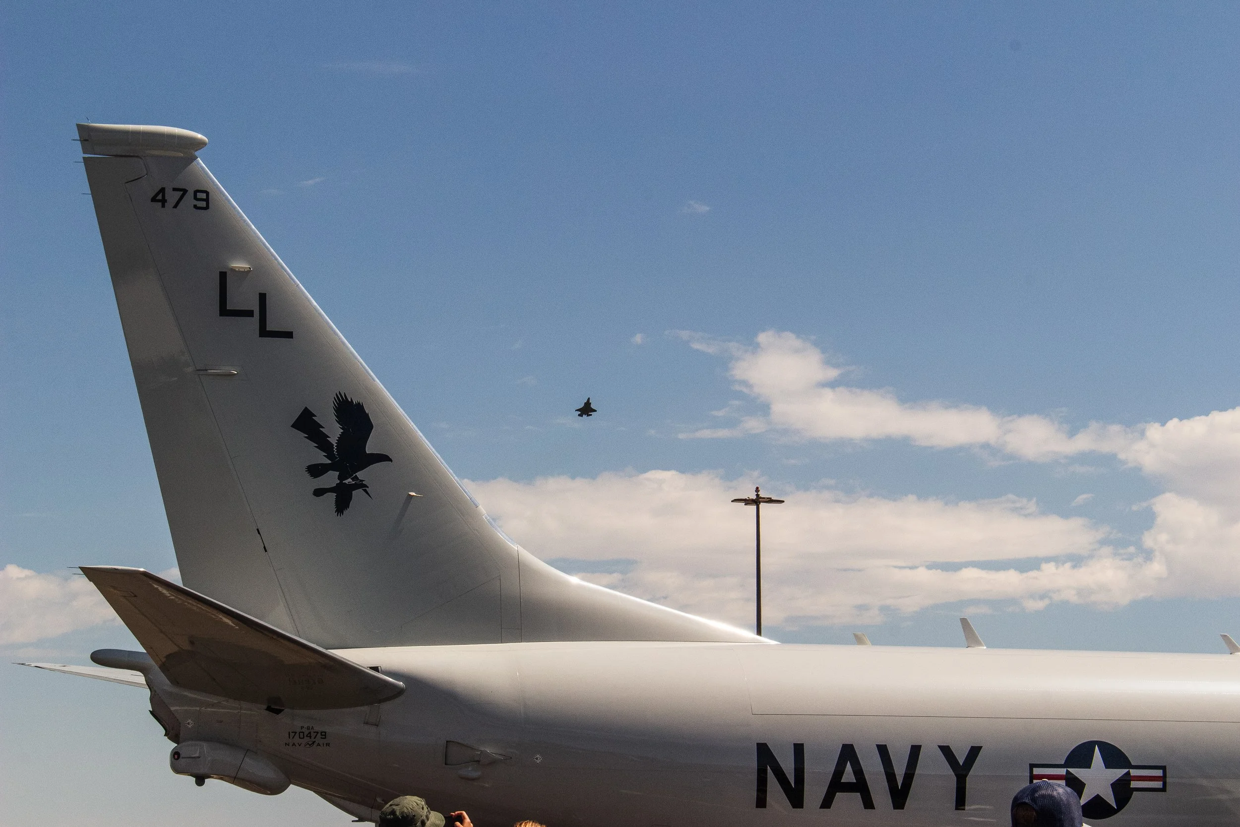 Close-up of the tail of a U.S. Navy aircraft with the letters 'LL' and the aircraft number '479' visible. The tail also features a black eagle logo. The background shows a blue sky with some clouds and another smaller jet flying in the distance.
