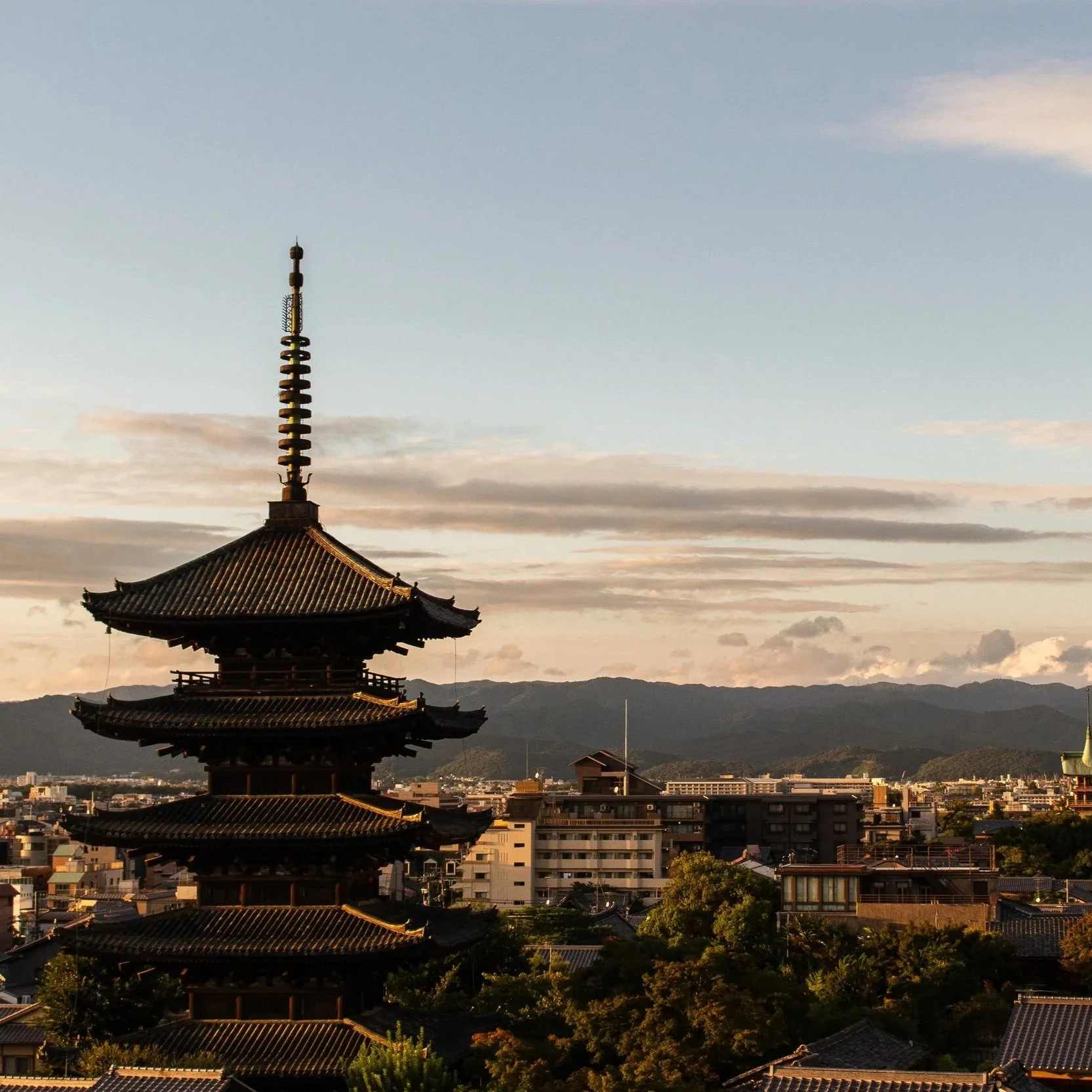 Traditional Japanese pagoda with a multi-tiered roof and a tall spire, set against a cityscape with modern buildings and distant mountains under a partly cloudy sky at sunset.