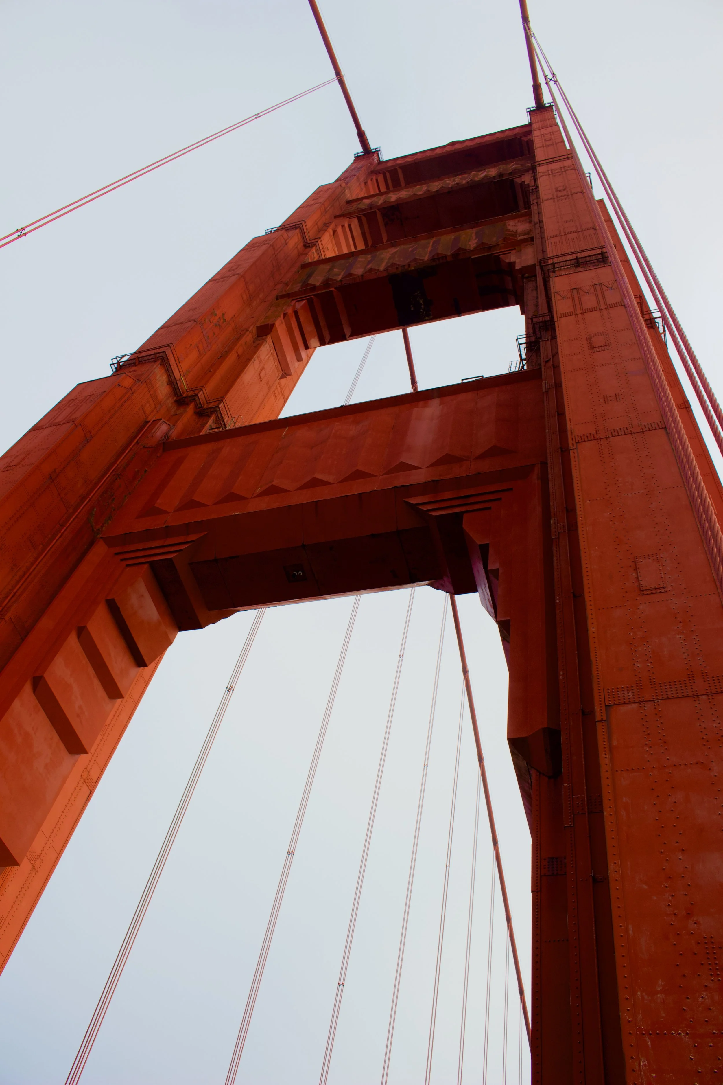 Close-up of the underside of a red-orange suspension bridge tower with cables extending from the top, against a clear sky.