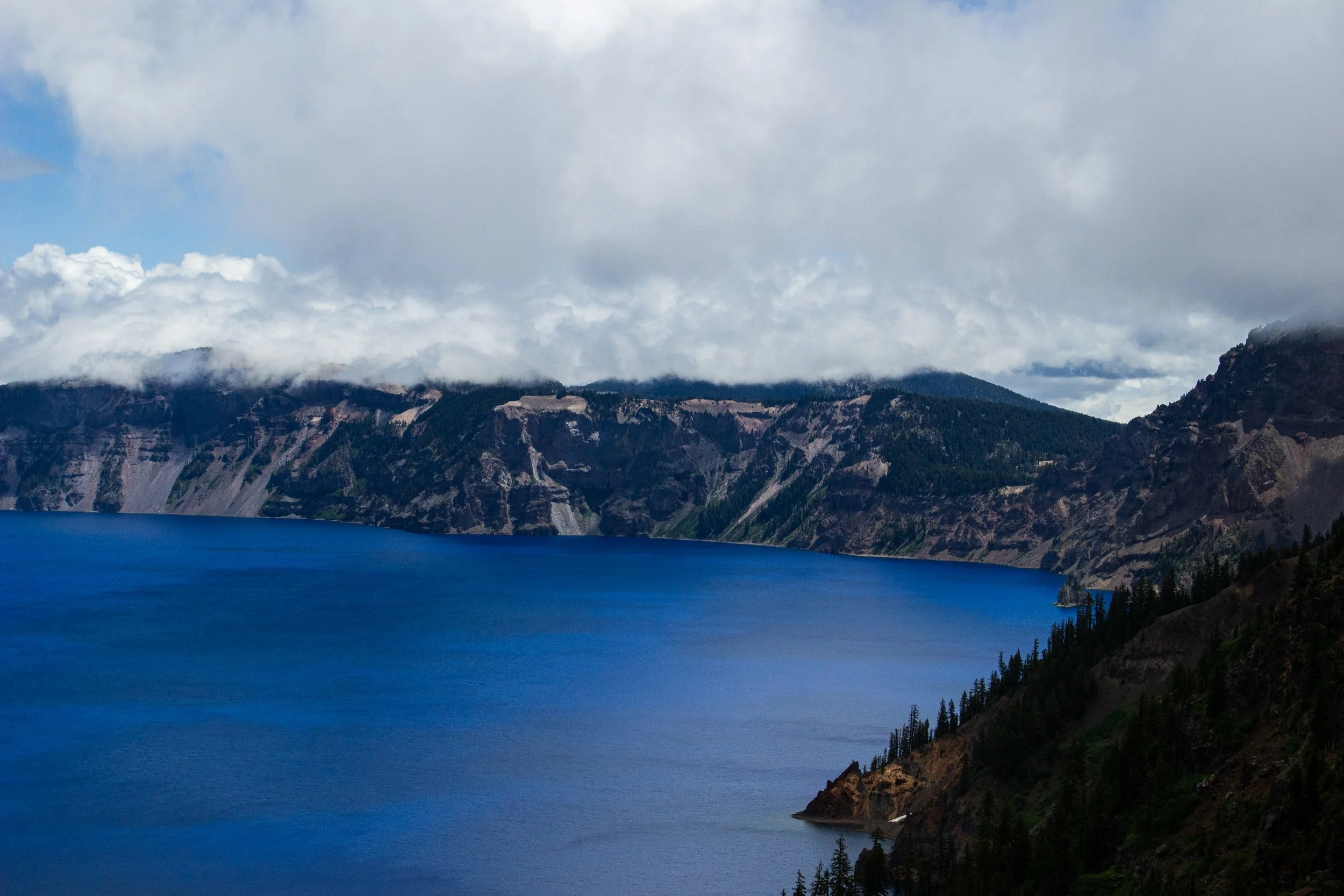 A large body of water with a deep blue color, surrounded by mountainous terrain with forests and rocky cliffs under a partly cloudy sky.