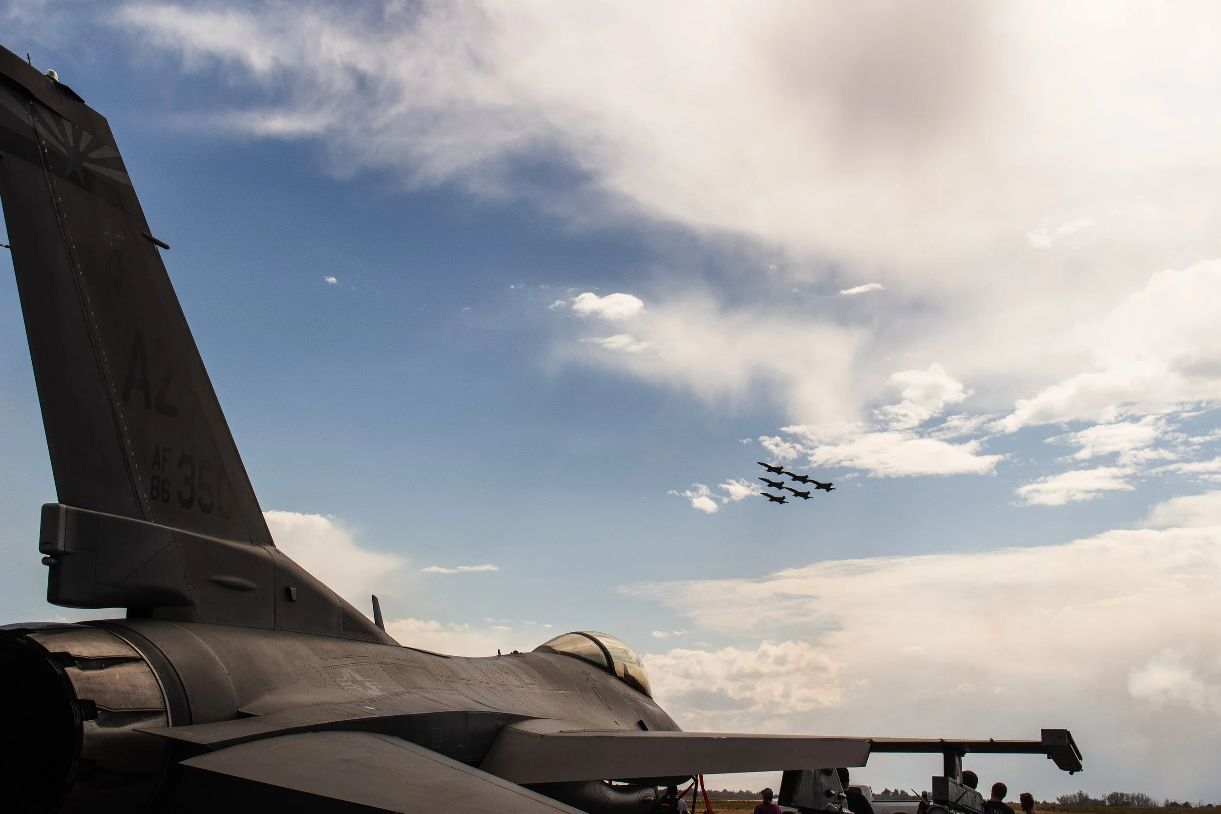 The image shows the tail section of a military jet fighter aircraft with some buildings and people in the background. The sky is partly cloudy with a formation of five fighter jets flying in the distance.