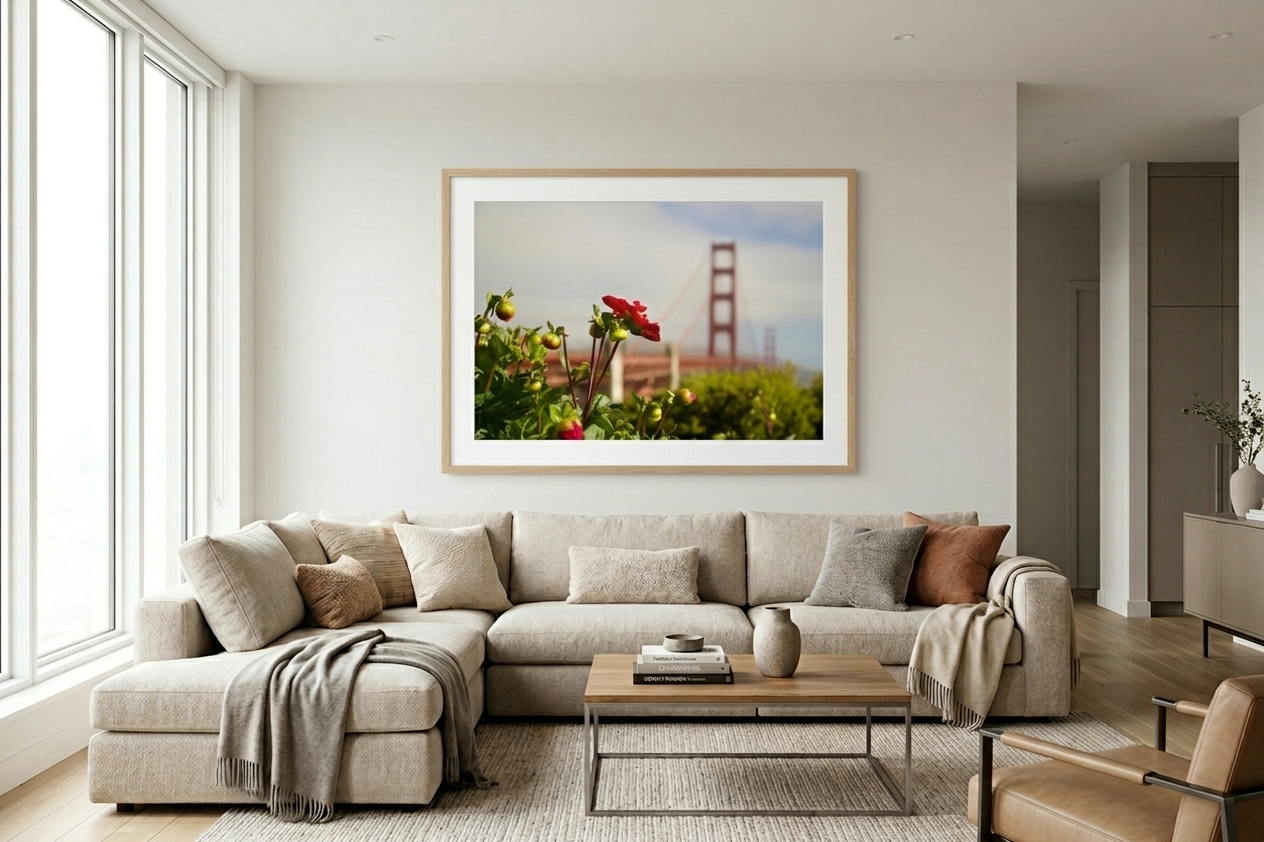 Living room with beige sectional sofa, cushions, a wooden coffee table with vases and books, large windows, and a framed photo of the Golden Gate Bridge with flowers in front.